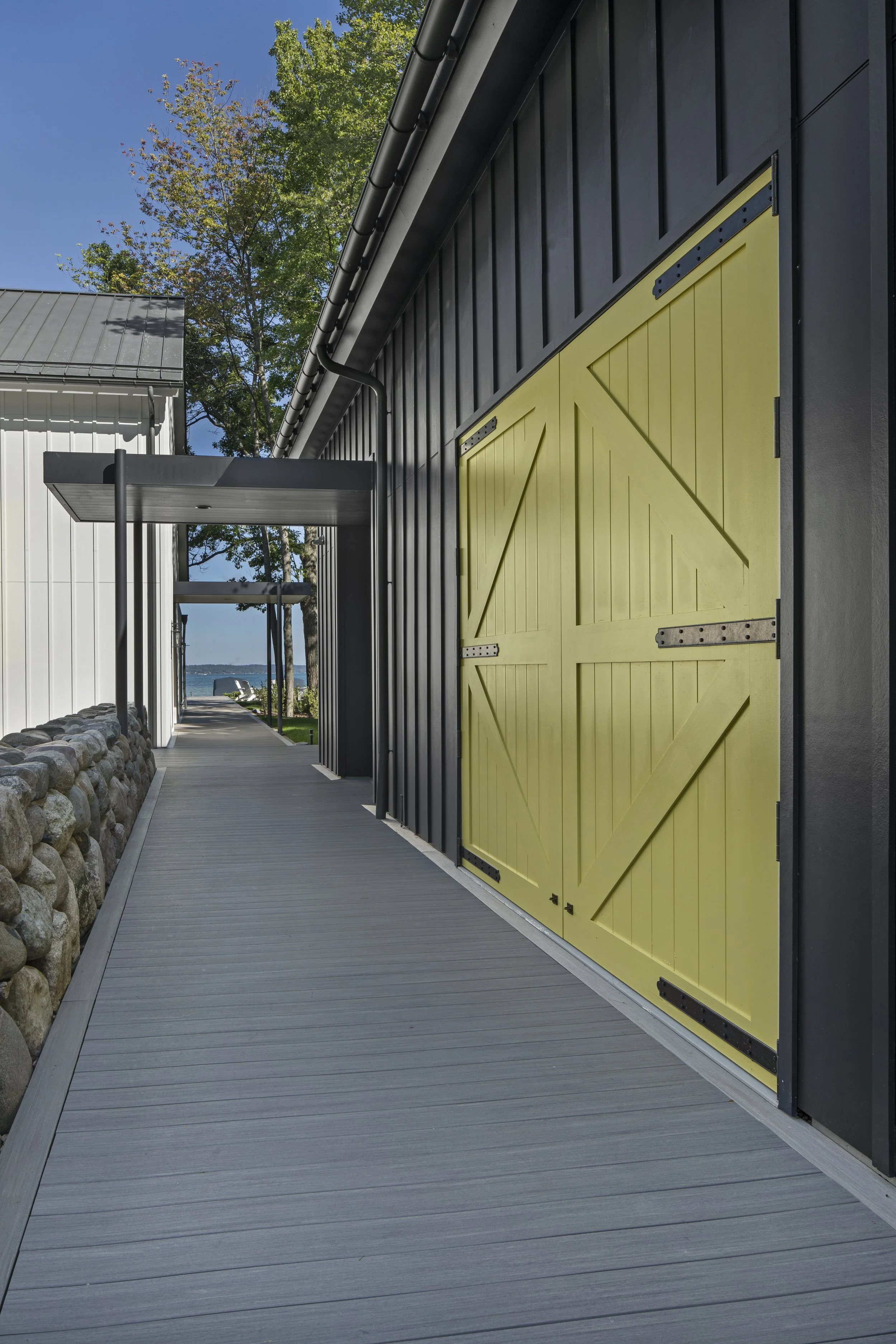 A modern barn-style building with black vertical siding and a yellow sliding barn door, along a wooden walkway. In the background, there are trees and a body of water visible through an archway.