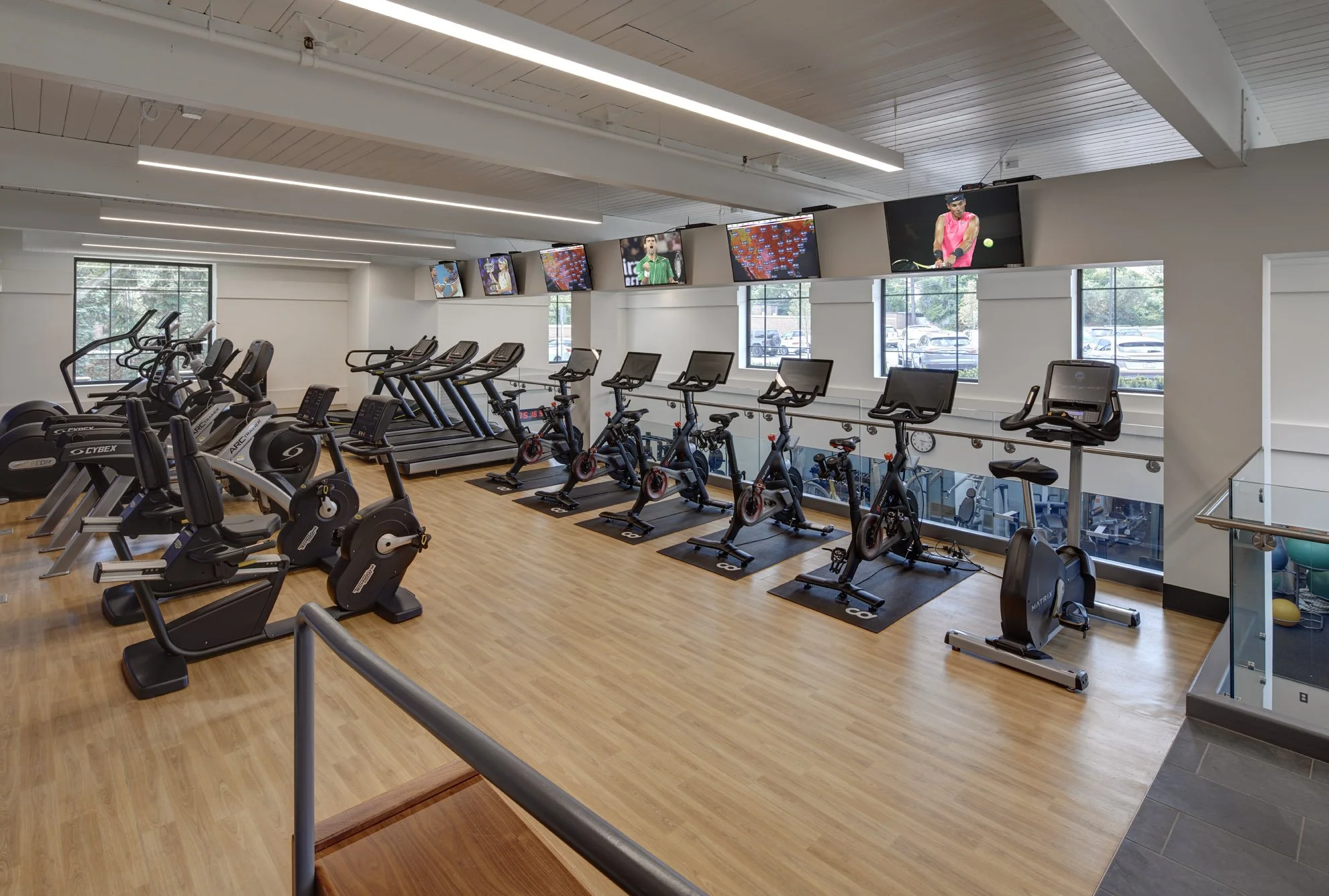 Inside a modern gym with cardio equipment including treadmills, stationary bikes, and elliptical machines, along with several wall-mounted TV screens showing various programming, large windows for natural light, and a wooden ceiling.