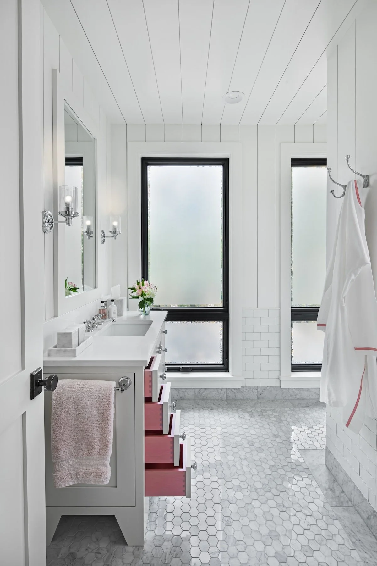 Bright and modern bathroom with white shiplap walls, hexagonal tile flooring, a white vanity with a pink drawer unit, and black-framed windows with frosted glass. There are wall-mounted light fixtures, a towel, and decorative flowers.