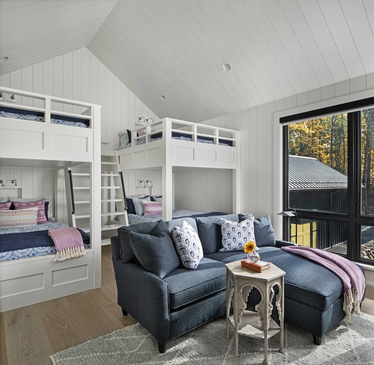 A cozy bedroom with a sloped ceiling, featuring two sets of white bunk beds and a dark blue sofa with patterned pillows. There is a small wooden side table with a sunflower in a vase and two books, placed in front of a large window showing a view of 