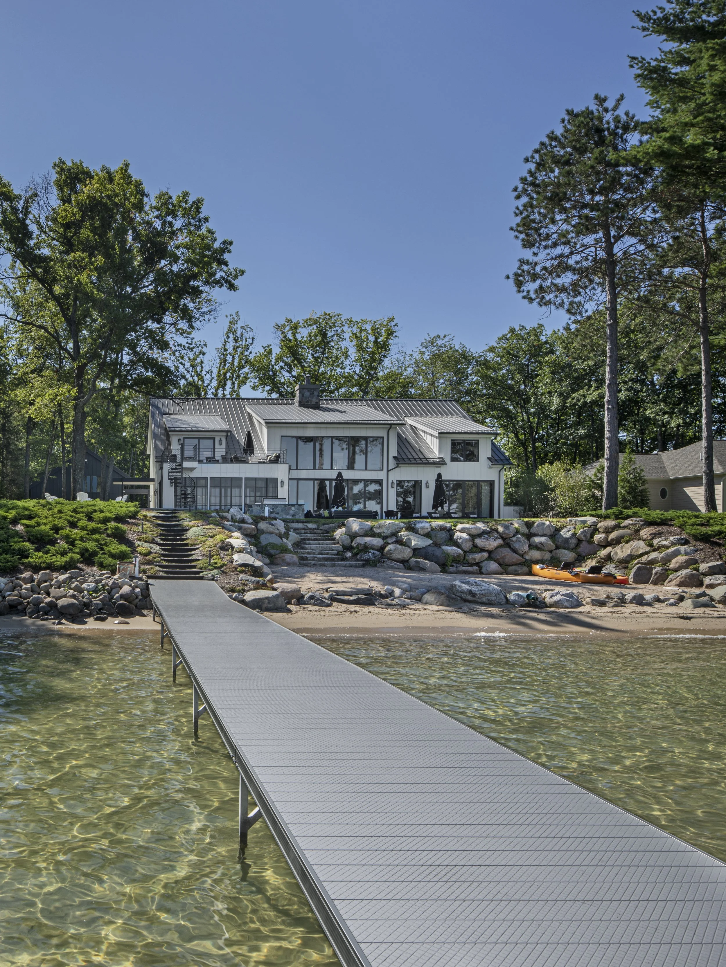 A dock extends from the water toward a house with a metal roof and large windows, surrounded by trees on a sunny day.