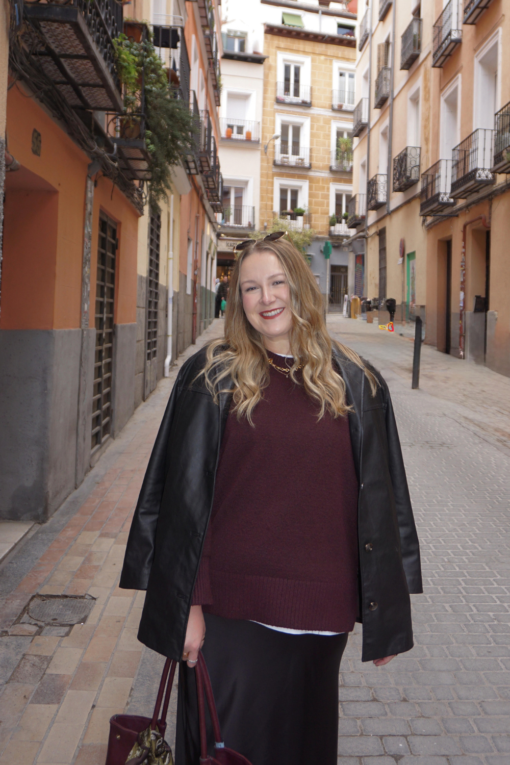 Young woman with long blonde hair smiling in a city street with colorful apartment buildings, wearing a black leather jacket, maroon sweater, and black skirt, holding a burgundy purse.