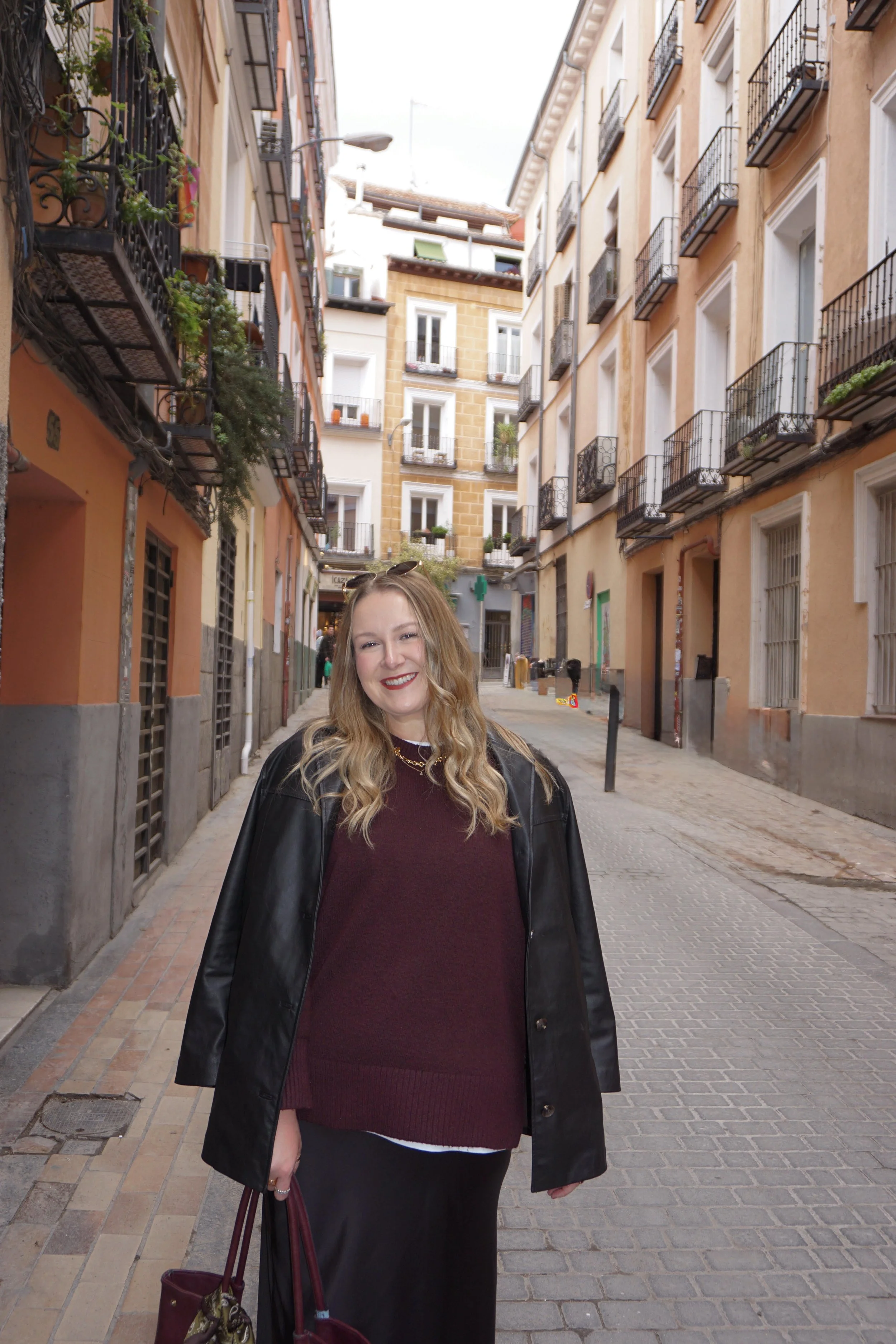 A smiling woman with blonde hair wearing a black leather jacket, maroon sweater, and black skirt, standing on a narrow city street with multi-story buildings and balconies.