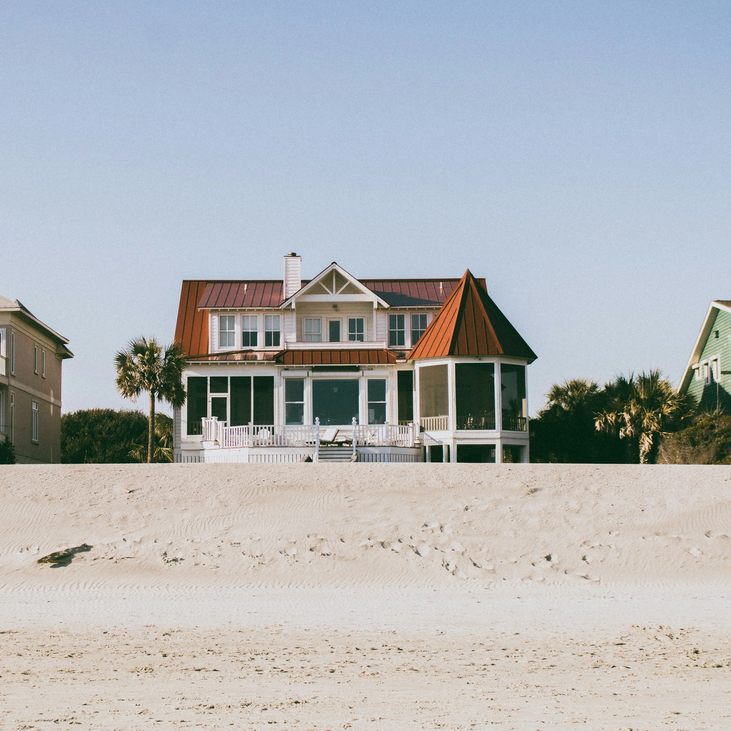 A large beach house with a red metal roof, white exterior, and multiple windows, situated behind sandy dunes and palm trees, under a clear blue sky.