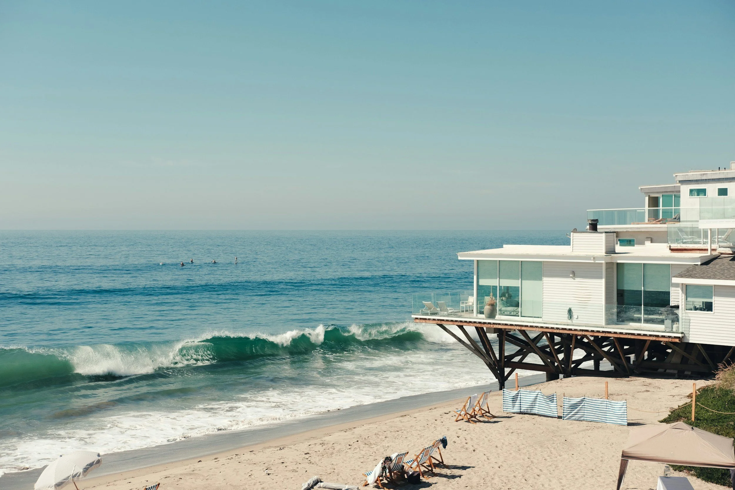Beach with a modern house on stilts facing the ocean, sandchairs on the beach, and waves approaching the shore under a clear sky.