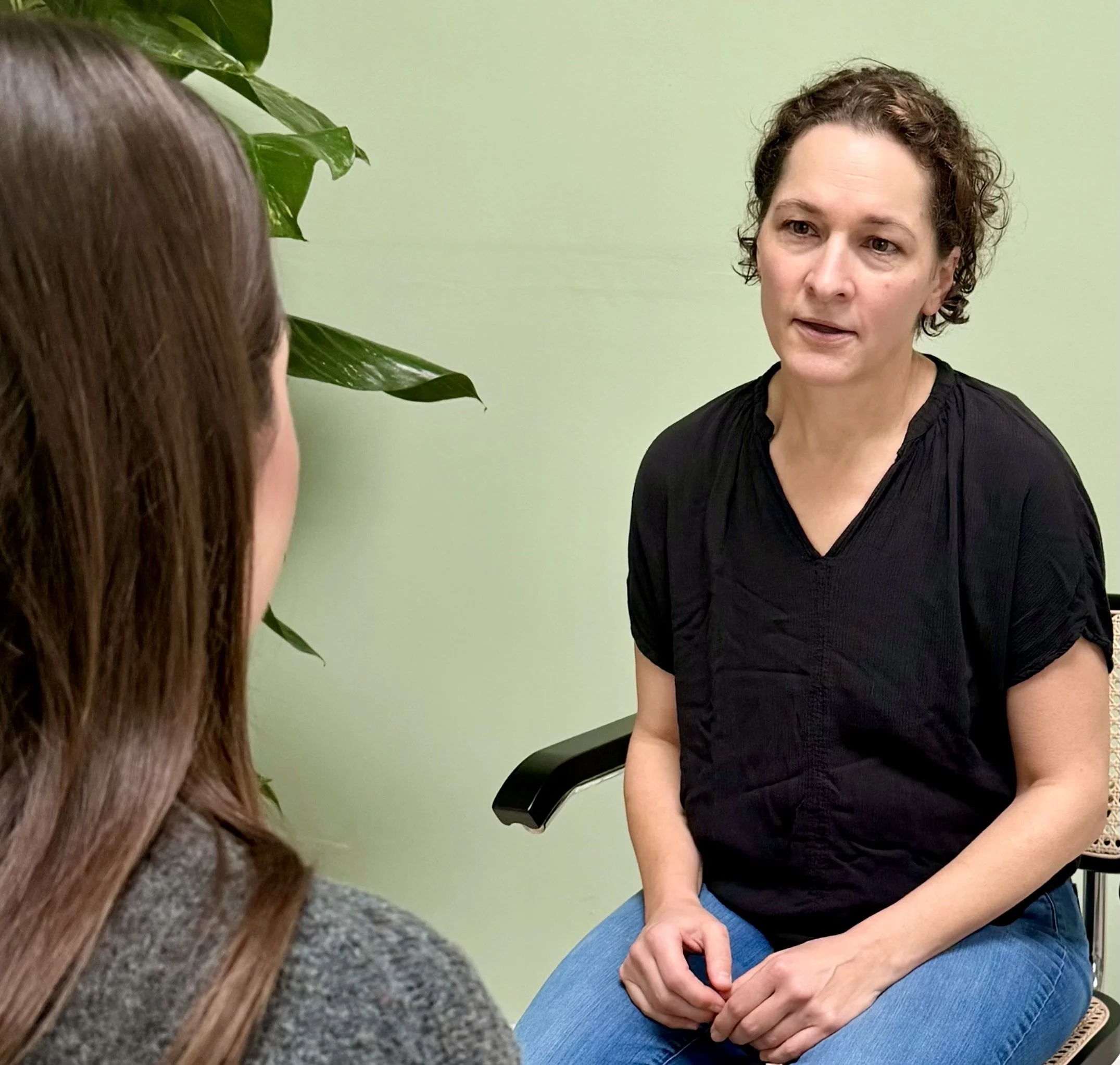 Zwei Frauen sprechen sich beim Sitzgespräch gegenübersitzend, eine mit dunklen, lockigen Haaren und die andere mit langen braunen Haaren, in einem Raum mit hellgrüner Wand und Pflanze im Hintergrund.