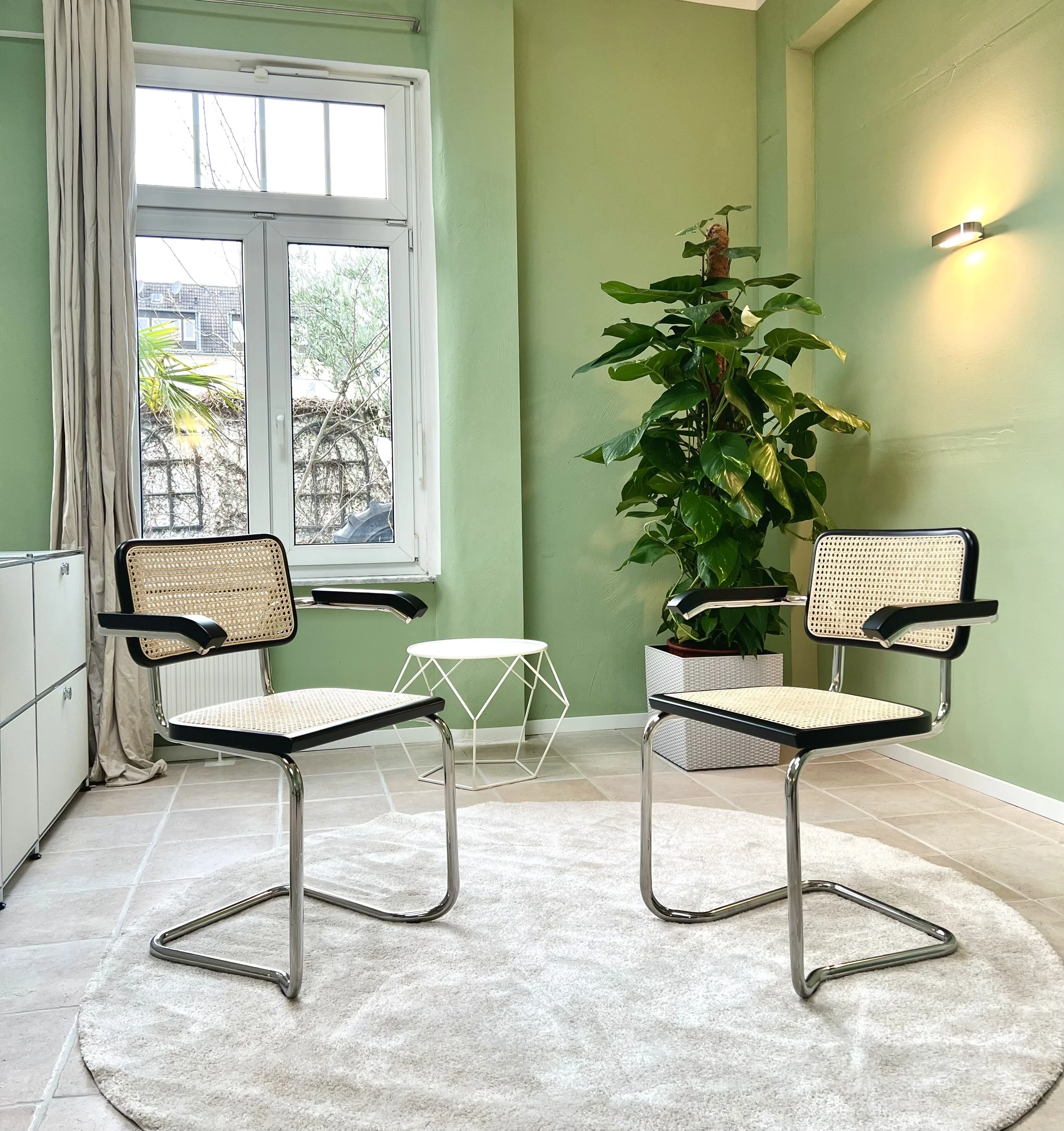 Ein gemütliches Wartezimmer mit zwei modernen Sessel, einem kleinen weißen Beistelltisch, einer grünen Wand, einer Zimmerpflanze in einer weißen Kiste, einem Fenster mit Blick auf Bäume und Häuser, einem Teppich auf Fliesenboden und einer Wandlampe.