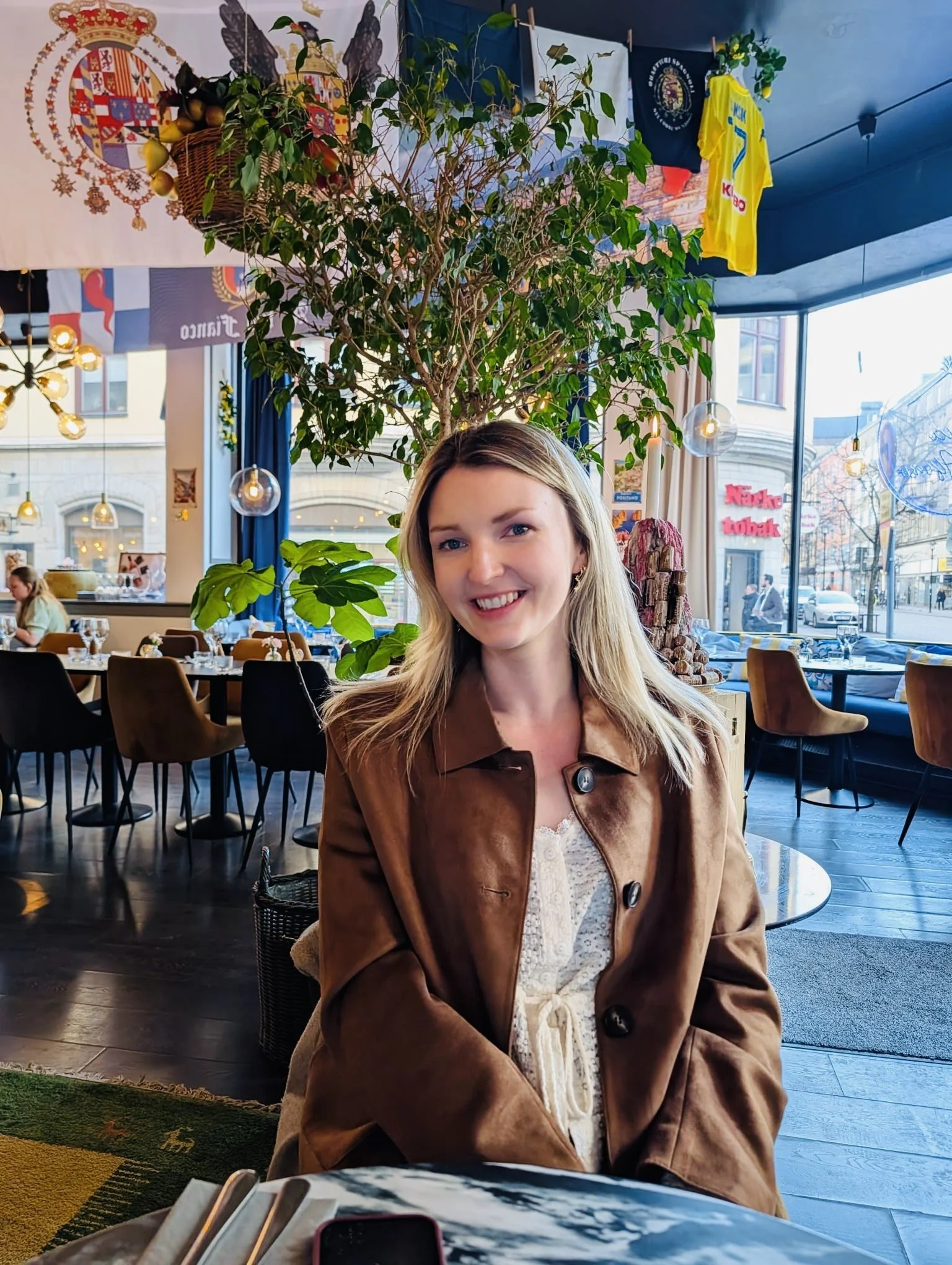A smiling woman with long blonde hair sitting in a restaurant with large windows, trees outside, and flags hanging from the ceiling, including a yellow sports jersey and various banners.