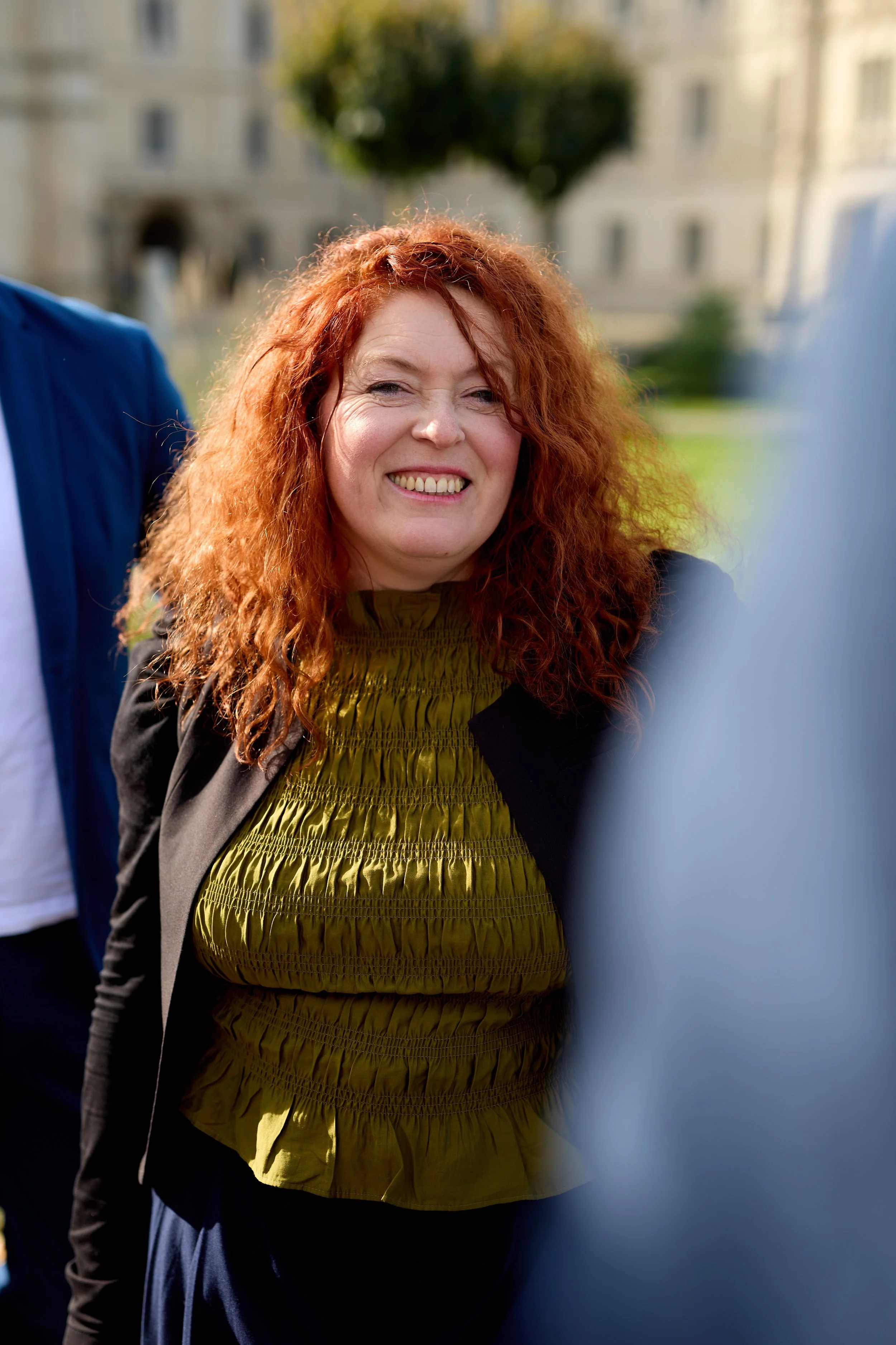 Fröhliche Frau mit lockigem roten Haar, lächelnd im Freien vor einer grünen Park- oder Gartenanlage.