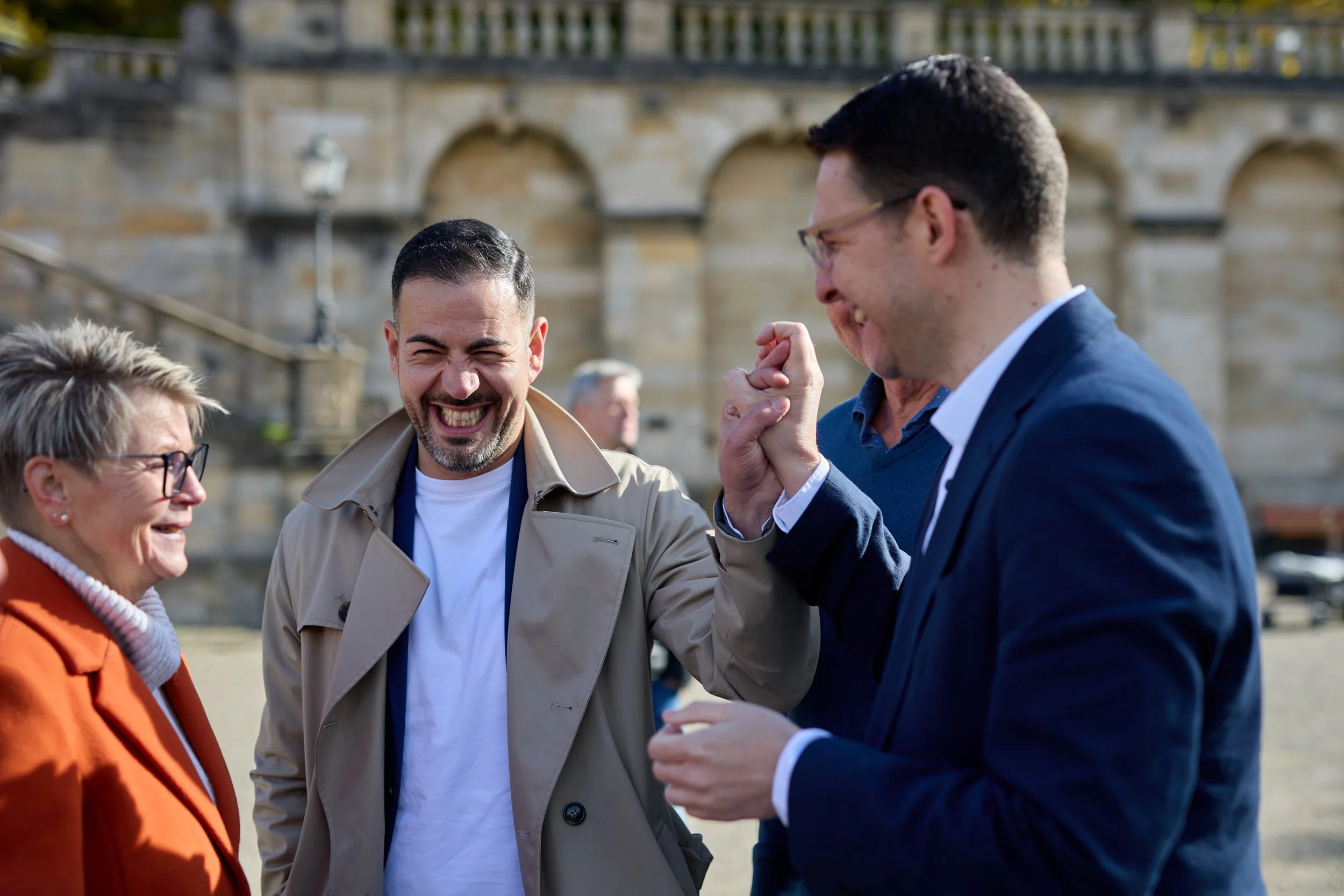 Vier Menschen lachen und unterhalten sich draußen vor einer alten Steinfassade, die vermutlich eine Brücke oder ein altes Gebäude ist.