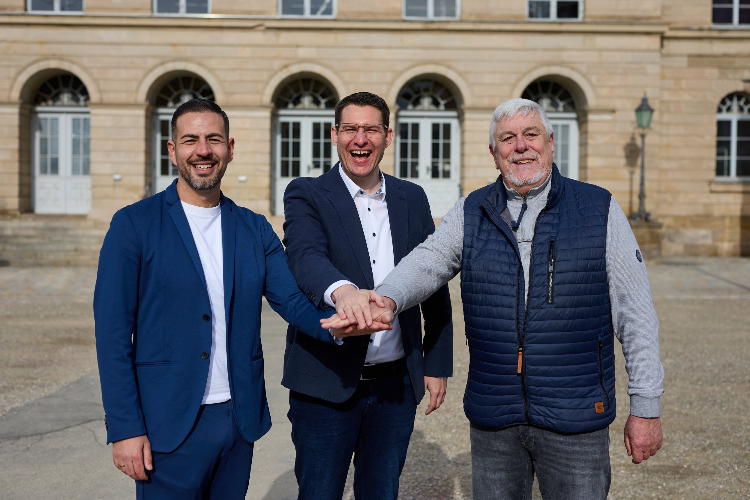 Drei Männer stehen vor einem historischen Gebäude, lachen und schütteln die Hände.