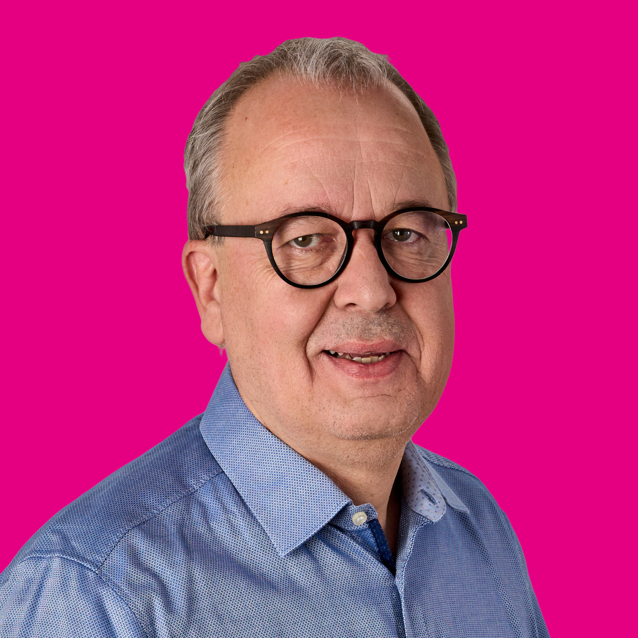 Ein Mann mit Kurzhaarschnitt, Brille und blauer Hemd vor pinkem Hintergrund.