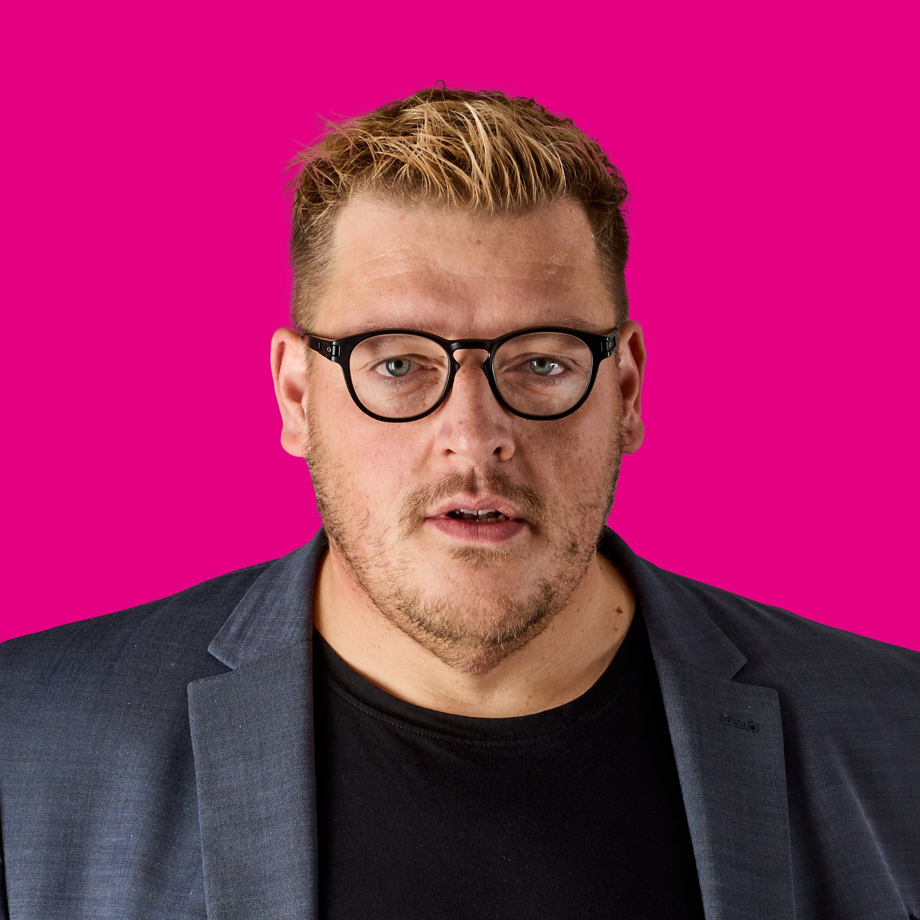 Junge Mann mit Brille, kurze blonde Haare, Kinnbart, trägt schwarze Jacke und schwarzes Shirt, vor pinkem Hintergrund.