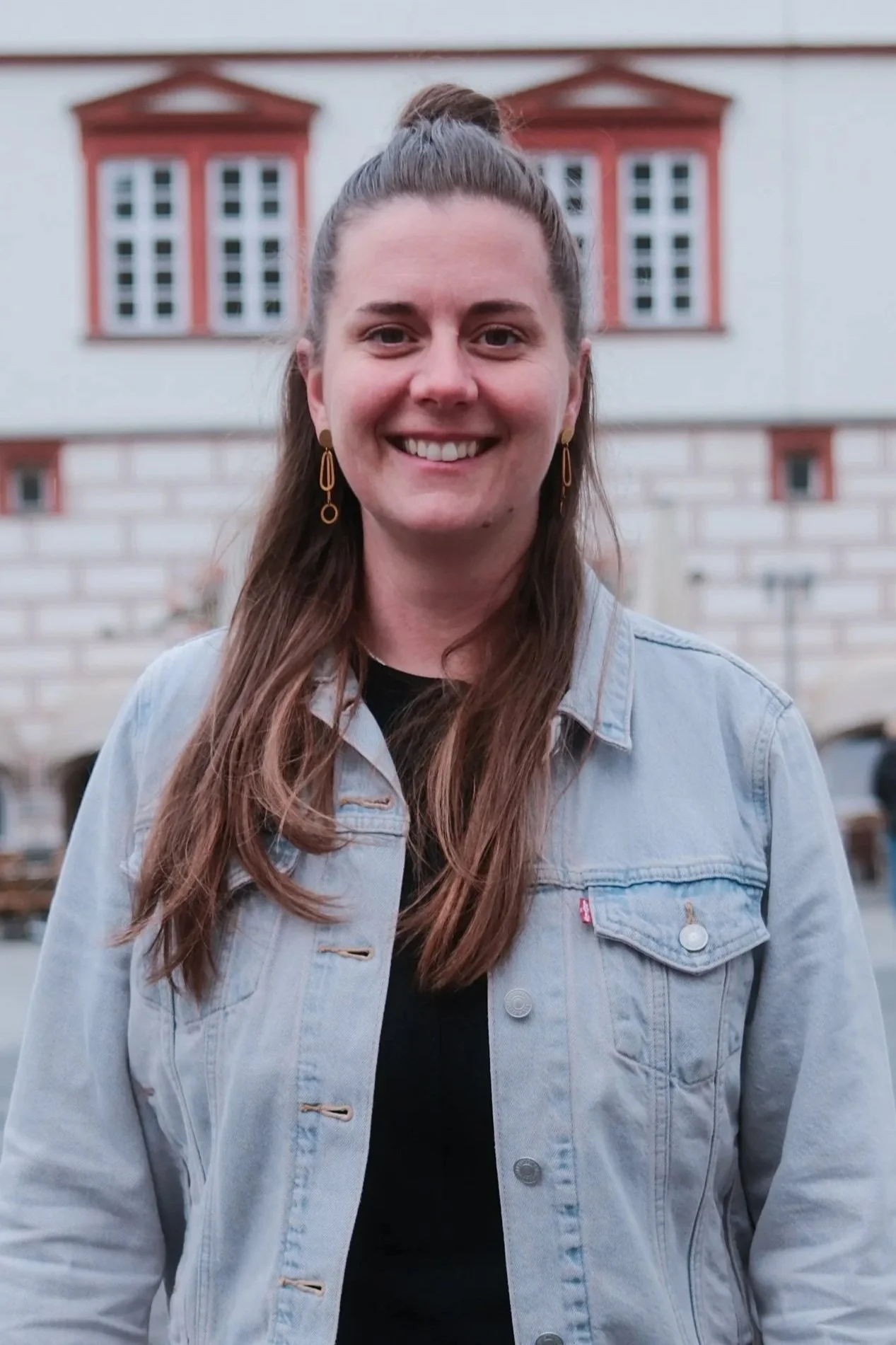 Porträt einer Frau mit braunen Haaren, die in eine Kamera blickt, trägt eine blaue Bluse, vor pinkem Hintergrund.