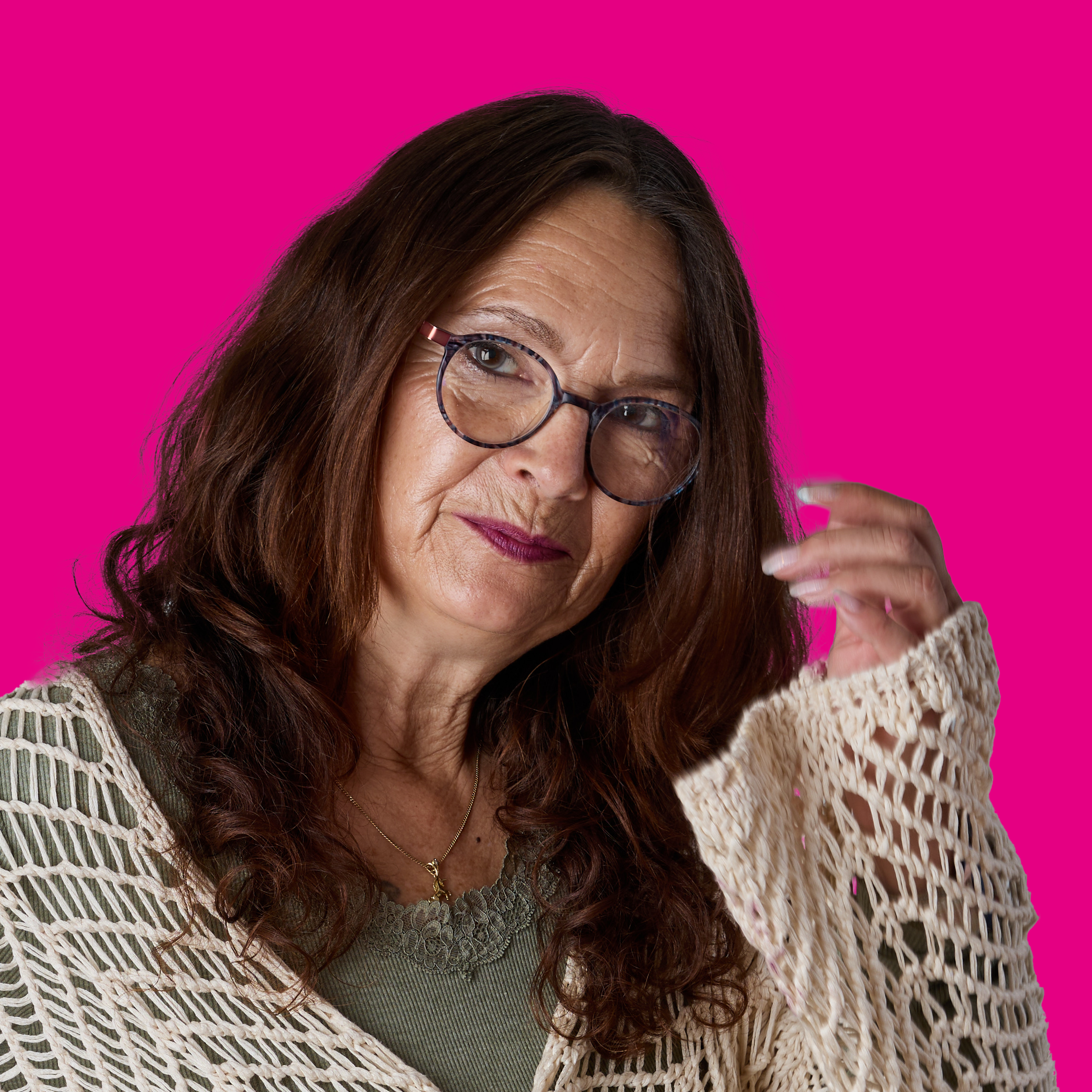 Eine ältere Frau mit braunen, lockigen Haaren und Brille, die an ihrer Seite hochgezogen wird, vor einem pinkfarbenen Hintergrund.