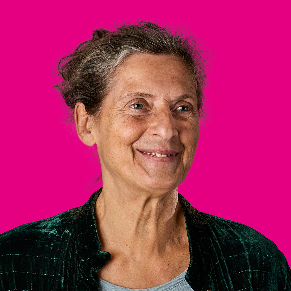 Porträt einer älteren Frau mit grauem Haar, lächelnd vor pinkem Hintergrund, trägt dunkle Jacke.
