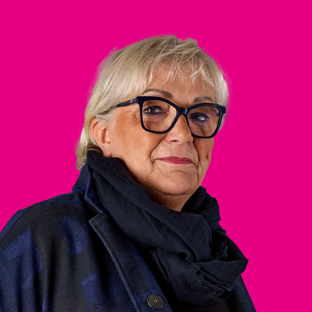 Porträt einer älteren Frau mit kurzen, grauen Haaren, schwarzen Brillen und dunkler Kleidung vor pinkem Hintergrund.