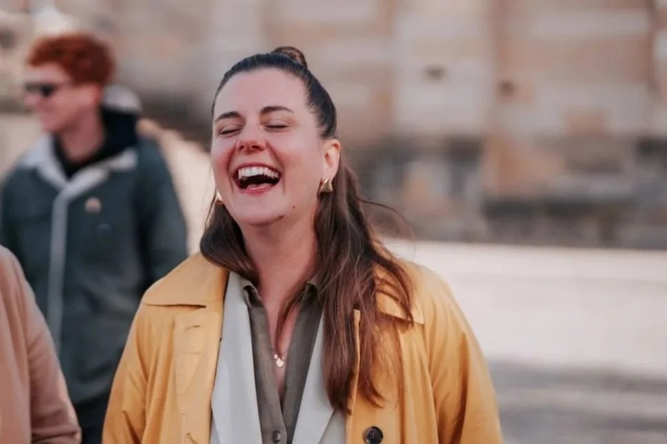 Eine lachende junge Frau mit braunen Haaren, die einen gelben Mantel trägt, steht im Freien vor einem Steinbau.