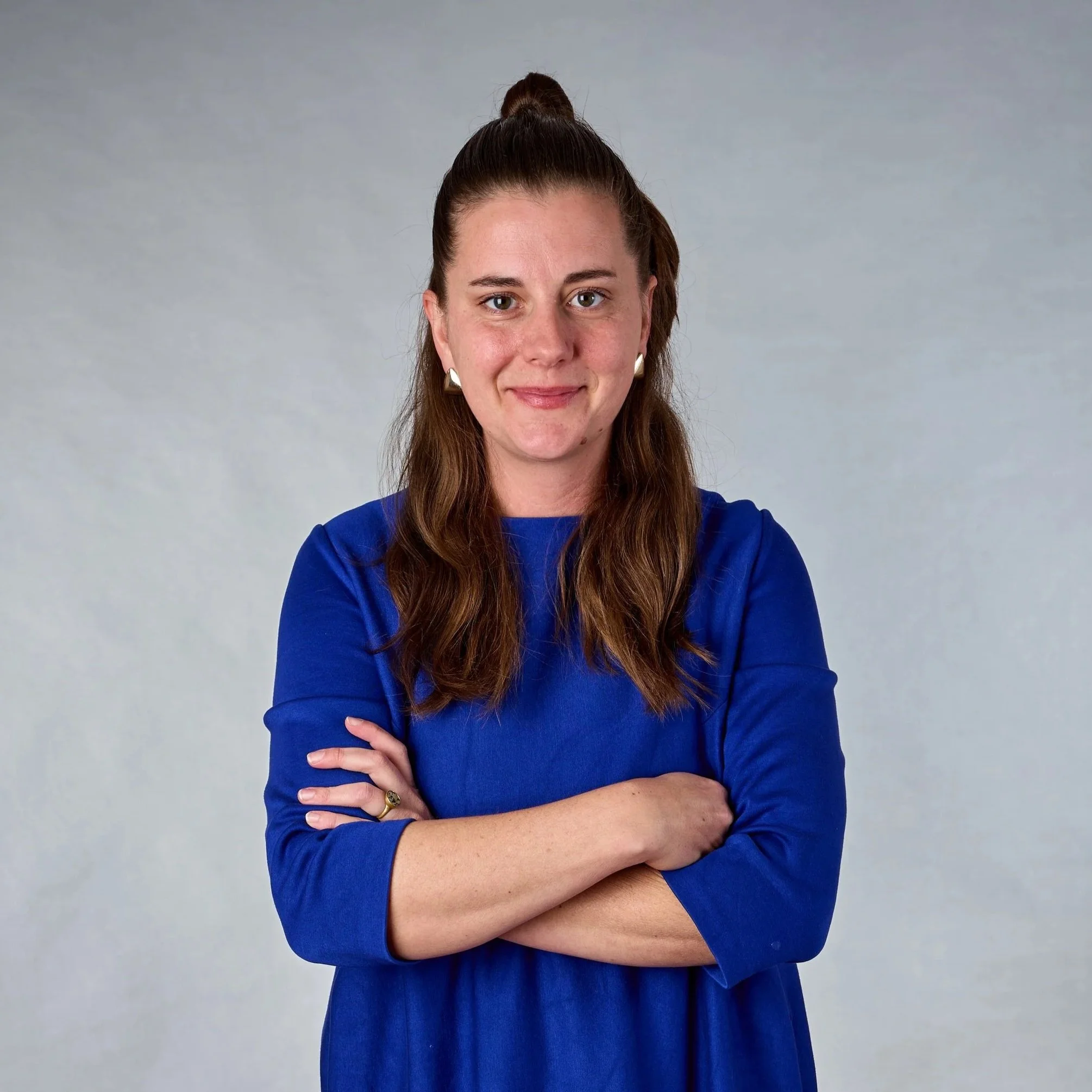 Junge Frau mit braunen Haaren, die einen Dutt trägt, trägt ein blaues Kleid, steht mit verschränkten Armen vor einem hellgrauen Hintergrund, lächelt leicht.