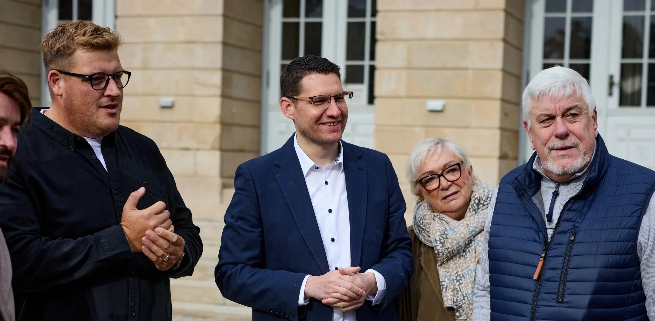 Eine Gruppe von fünf Menschen, bestehend aus drei Männern und zwei Frauen, steht vor einem gelben Ziegelgebäude mit weißen Fenstern. Sie scheinen sich im Gespräch zu befinden.