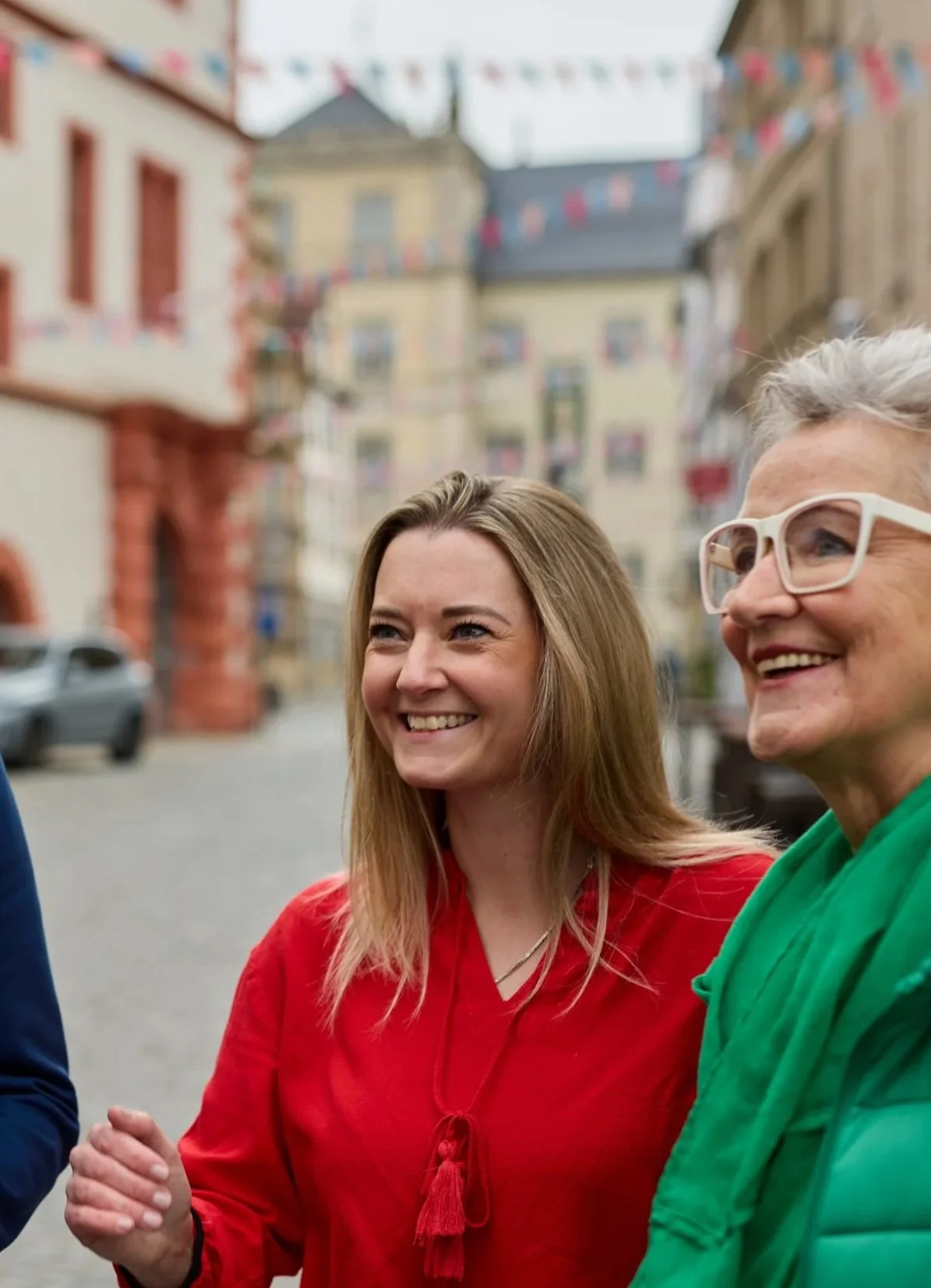 Zwei lachende Frauen, eine mit braunen Haaren in rotem Oberteil und die andere mit grauen Haaren in einer grünen Jacke, stehen im Freien vor bunten Häusern und bunter Dekoration.