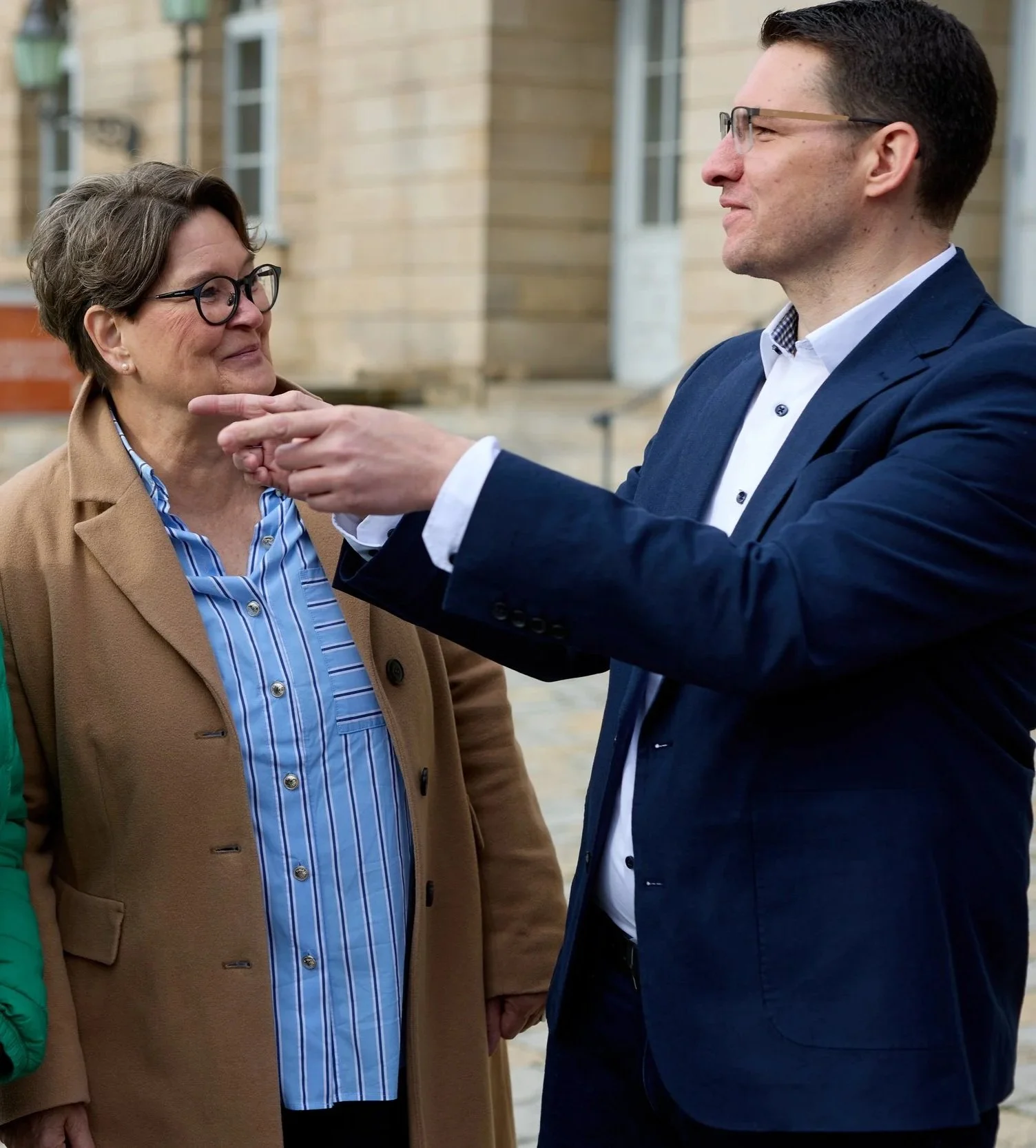 Ein Mann in einem dunklen Anzug und Brille lächelt und berührt eine ältere Frau im Kontrast, die ebenfalls Brille trägt und einen braunen Mantel trägt.