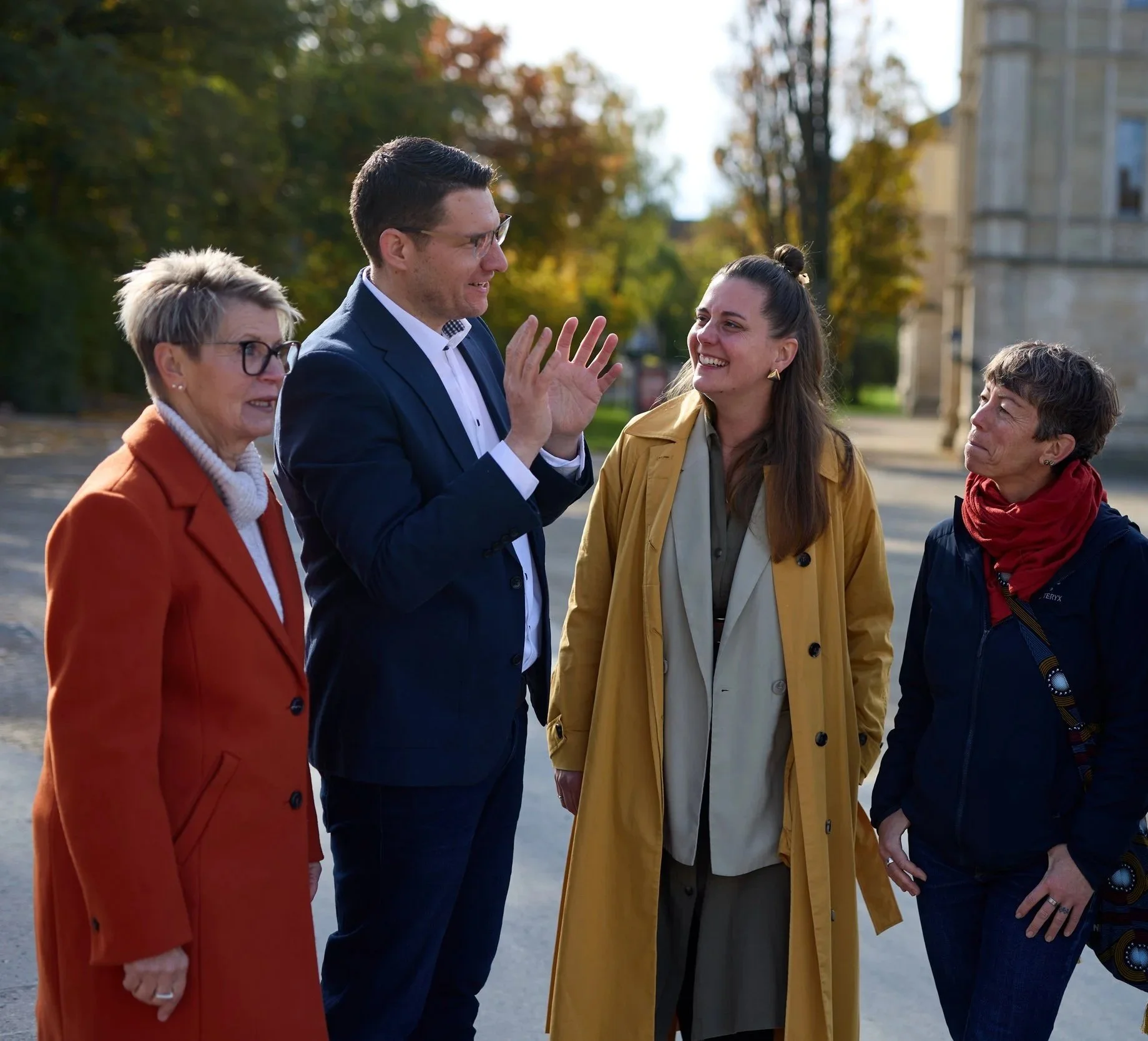 Vier Menschen im Gespräch, lachend und lächelnd, im Freien mit Herbstlaub und Gebäuden im Hintergrund.