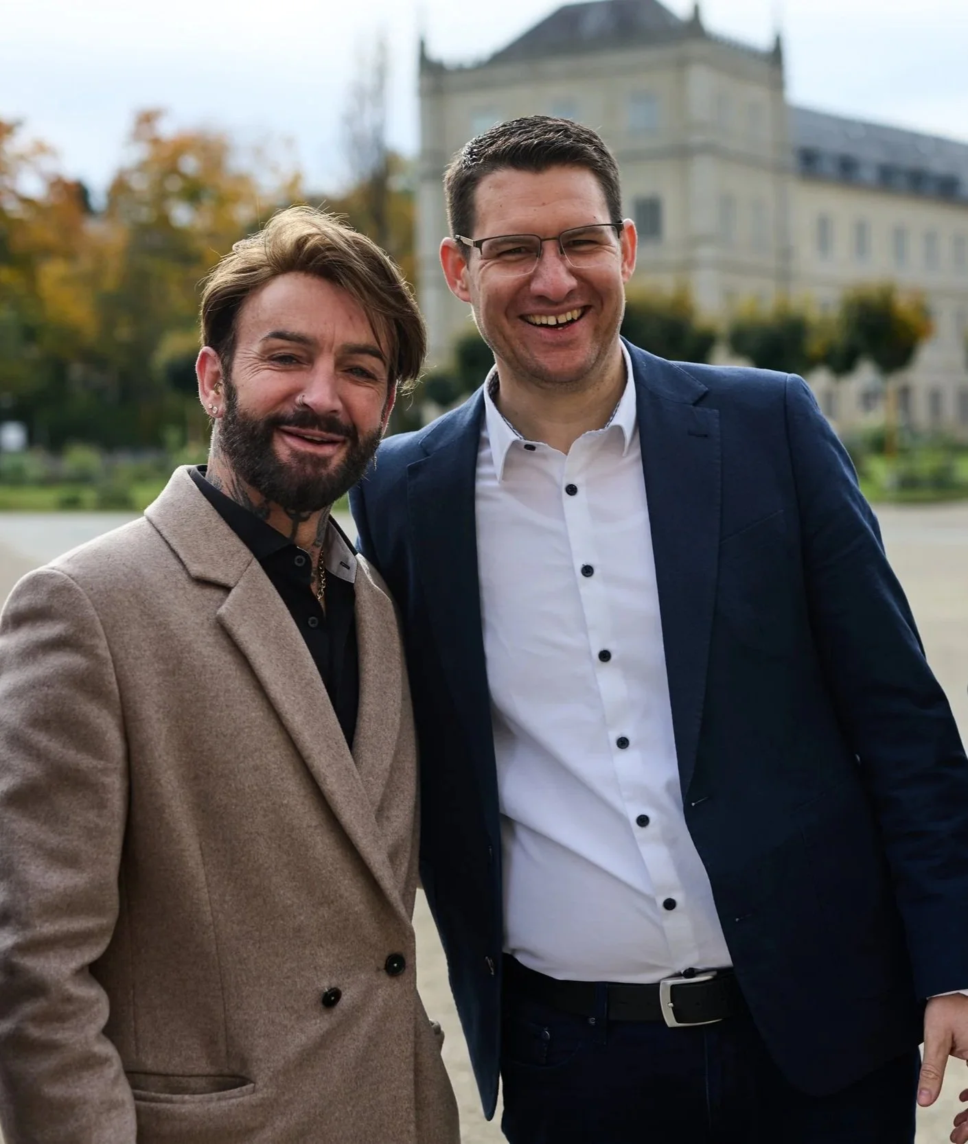 Zwei Männer in formeller Kleidung stehen draußen vor einem historischen Gebäude und lächeln in die Kamera.