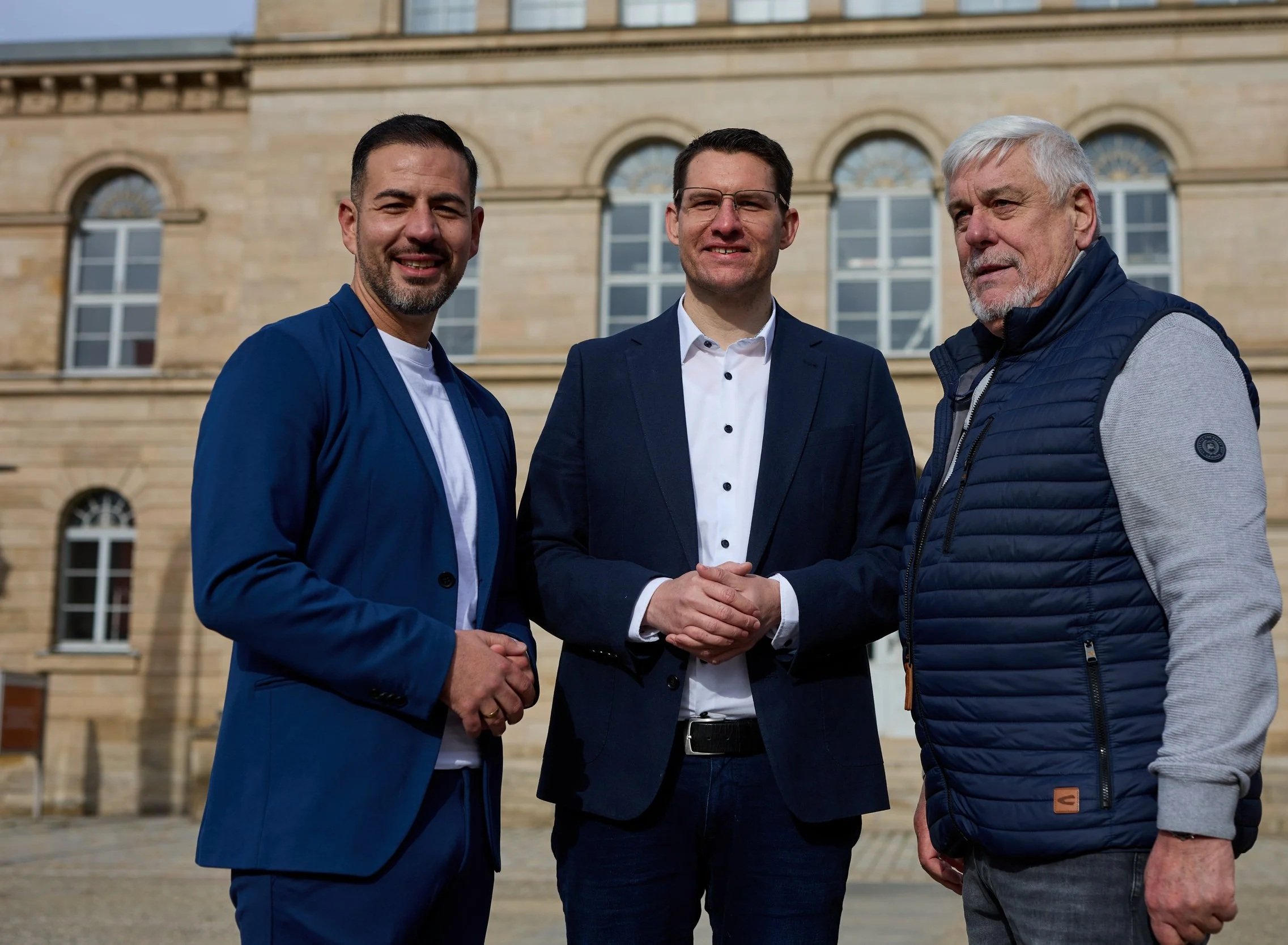 Drei Männer stehen vor einem historischen Gebäude mit großen Fenstern. Sie tragen formelle und halbformelle Kleidung und grüßen die Kamera.