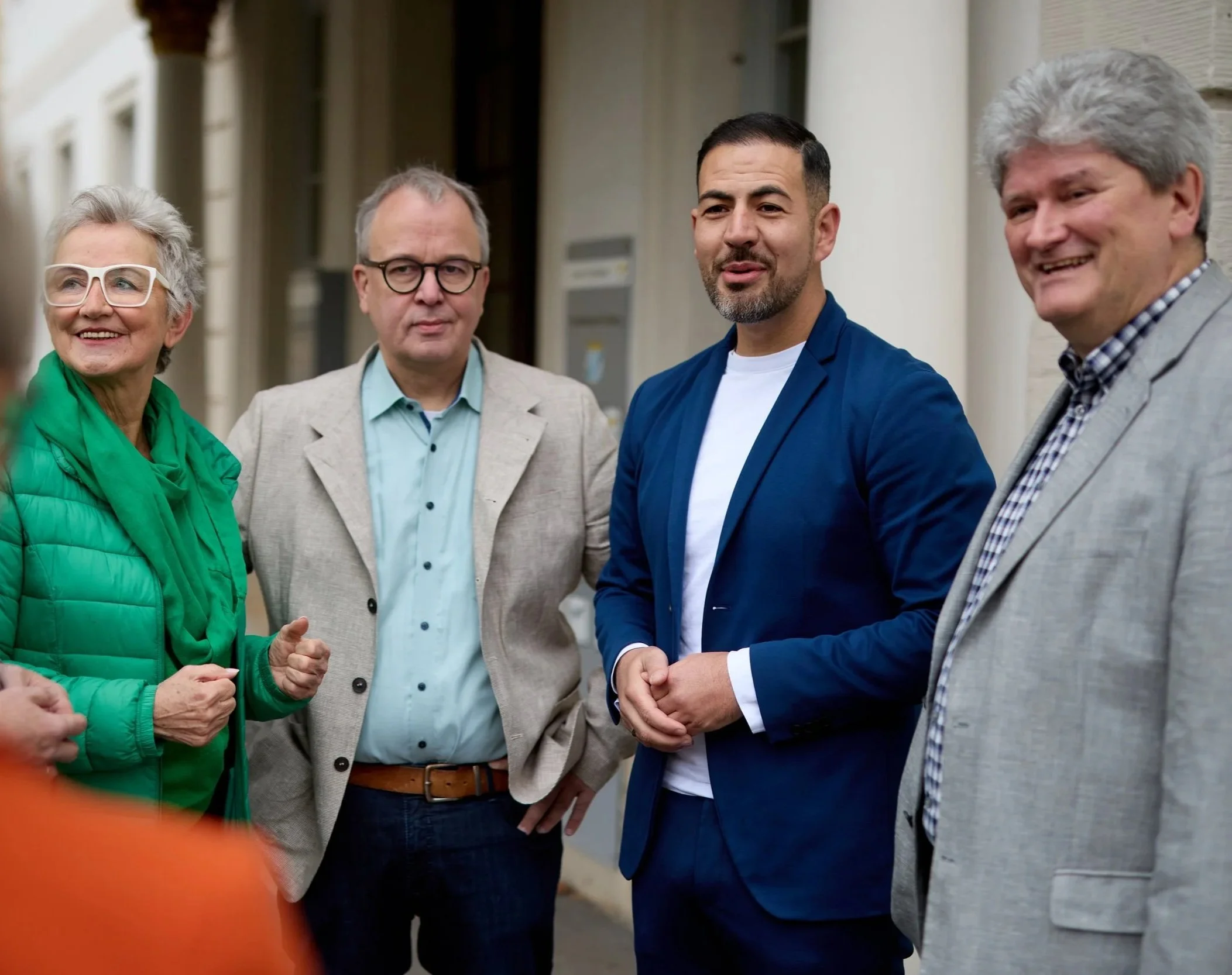 Fünf Menschen stehen draußen vor einem Haus. Sie sprechen miteinander und lachen, tragen formelle Kleidung. Es sind zwei Männer, zwei Frauen und ein weiterer Mann.