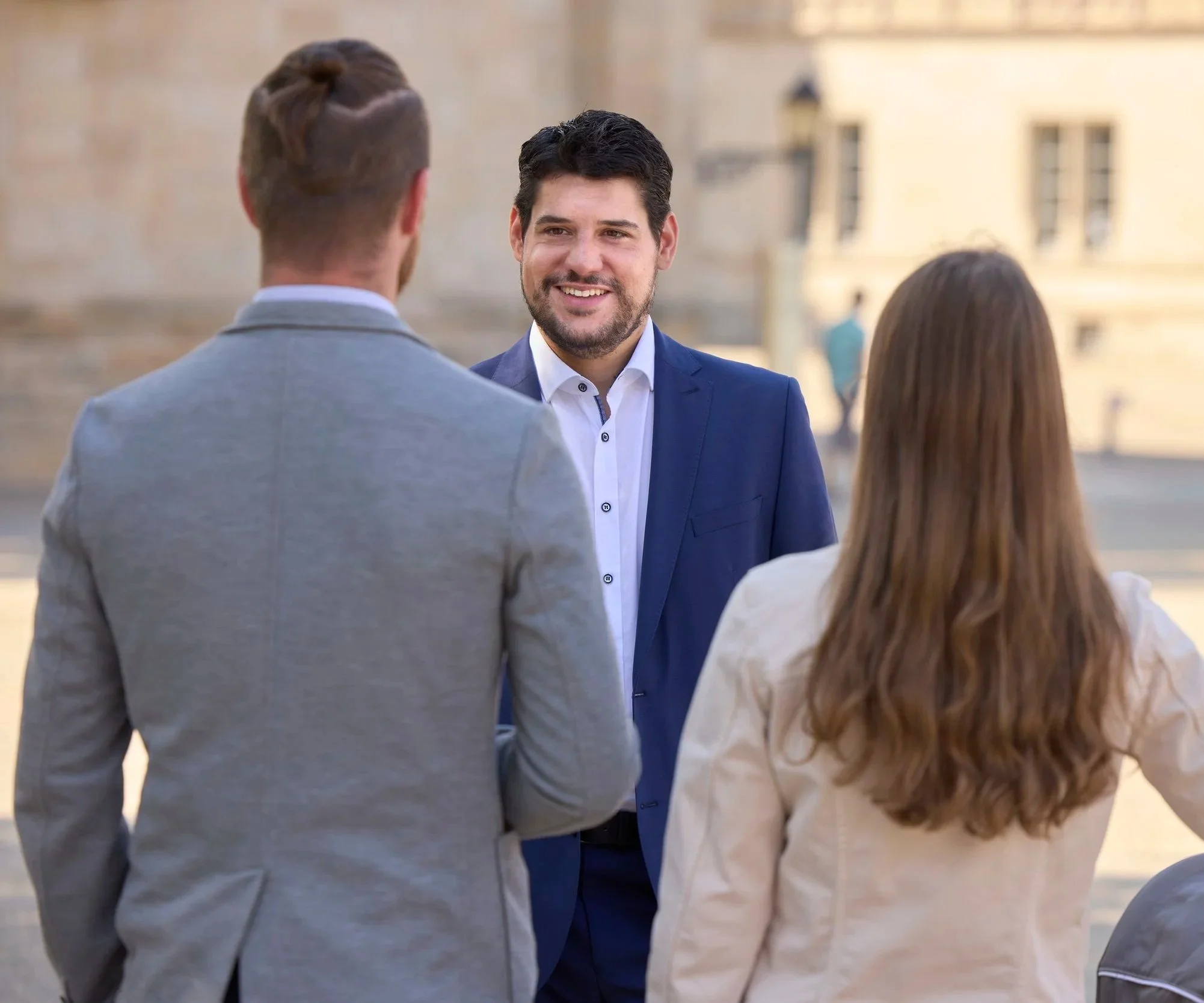 Drei Personen im Gespräch, zwei Männer und eine Frau, im Freien, in formeller Kleidung, lächelnd.