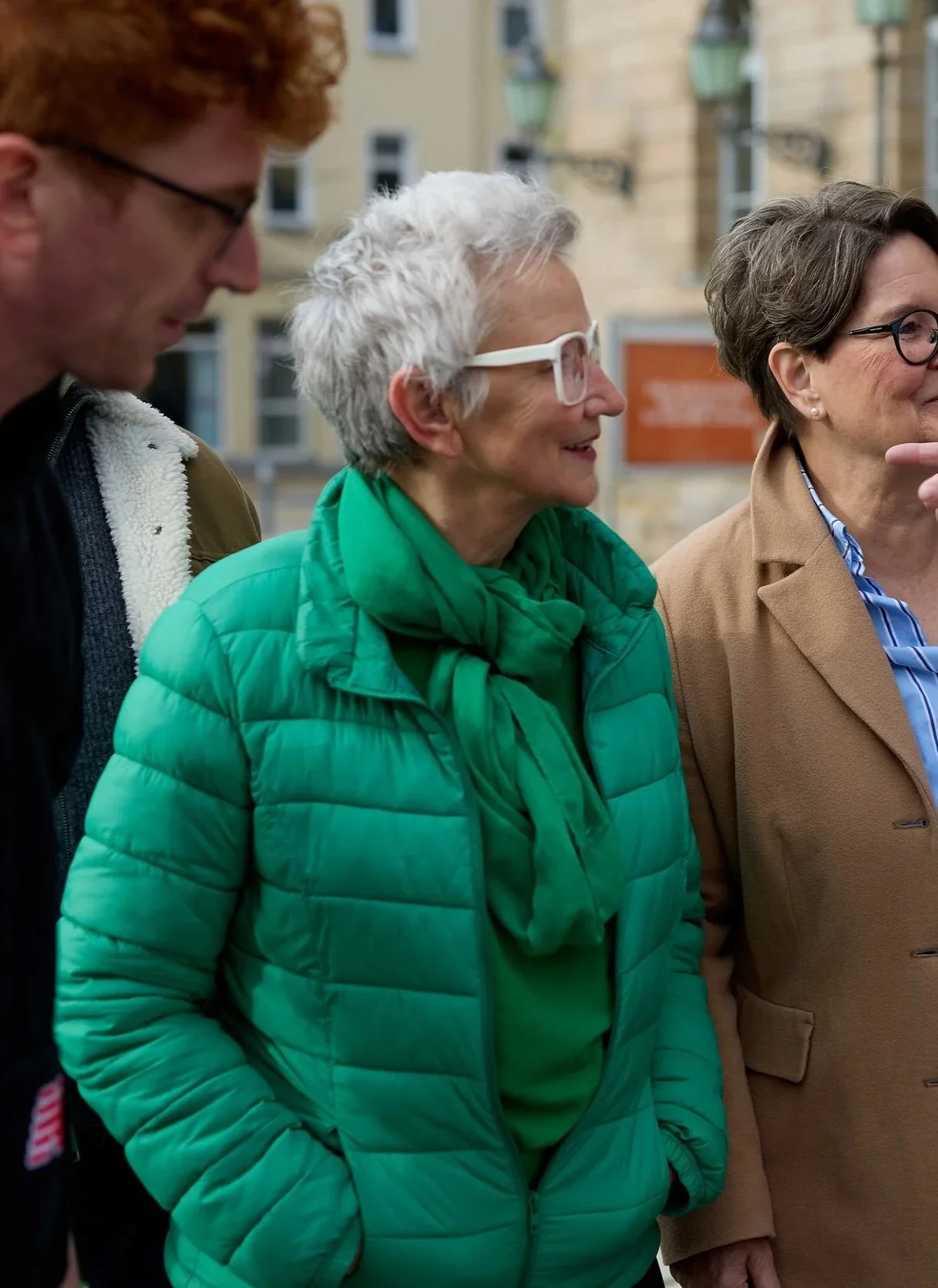 Drei Frauen und ein Mann stehen im Freien und führen ein Gespräch. Im Hintergrund sind Gebäude zu sehen.