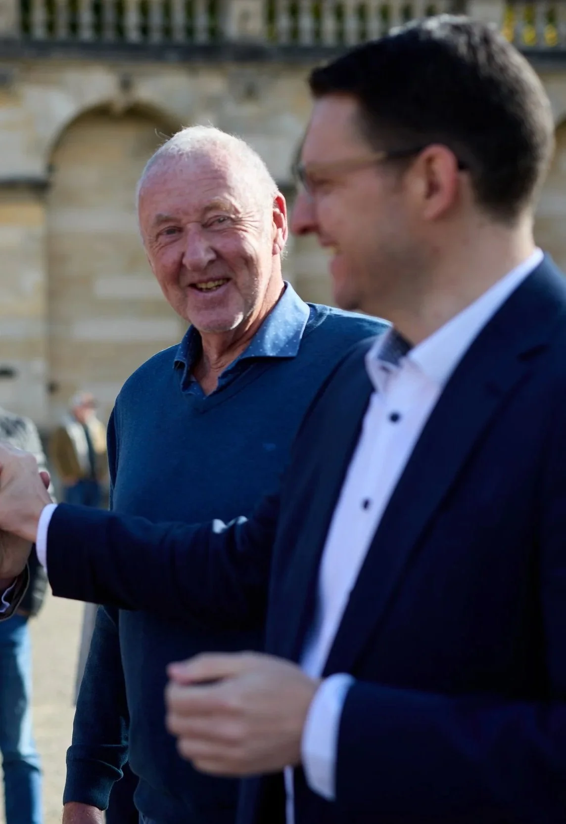 Zwei Männer im Gespräch und Lachen im Freien, eine Person in einem blauen Pullover, die andere in einem dunklen Anzug und weißen Hemd, im Hintergrund eine alte Steinfassade.