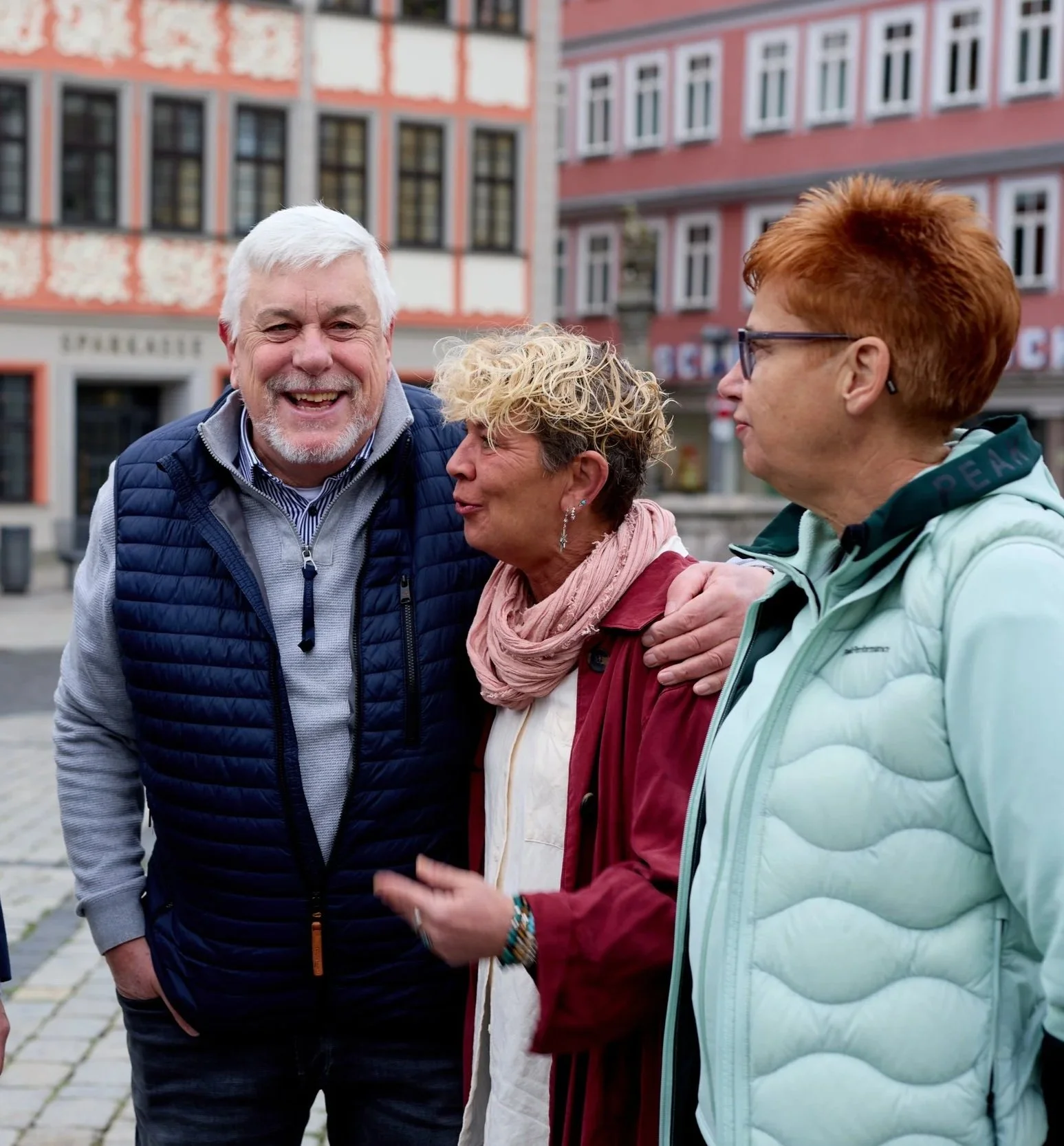 Drei ältere Menschen, zwei Frauen und ein Mann, unterhalten sich auf einer Straße in einer Stadt mit farbigen Häusern im Hintergrund.