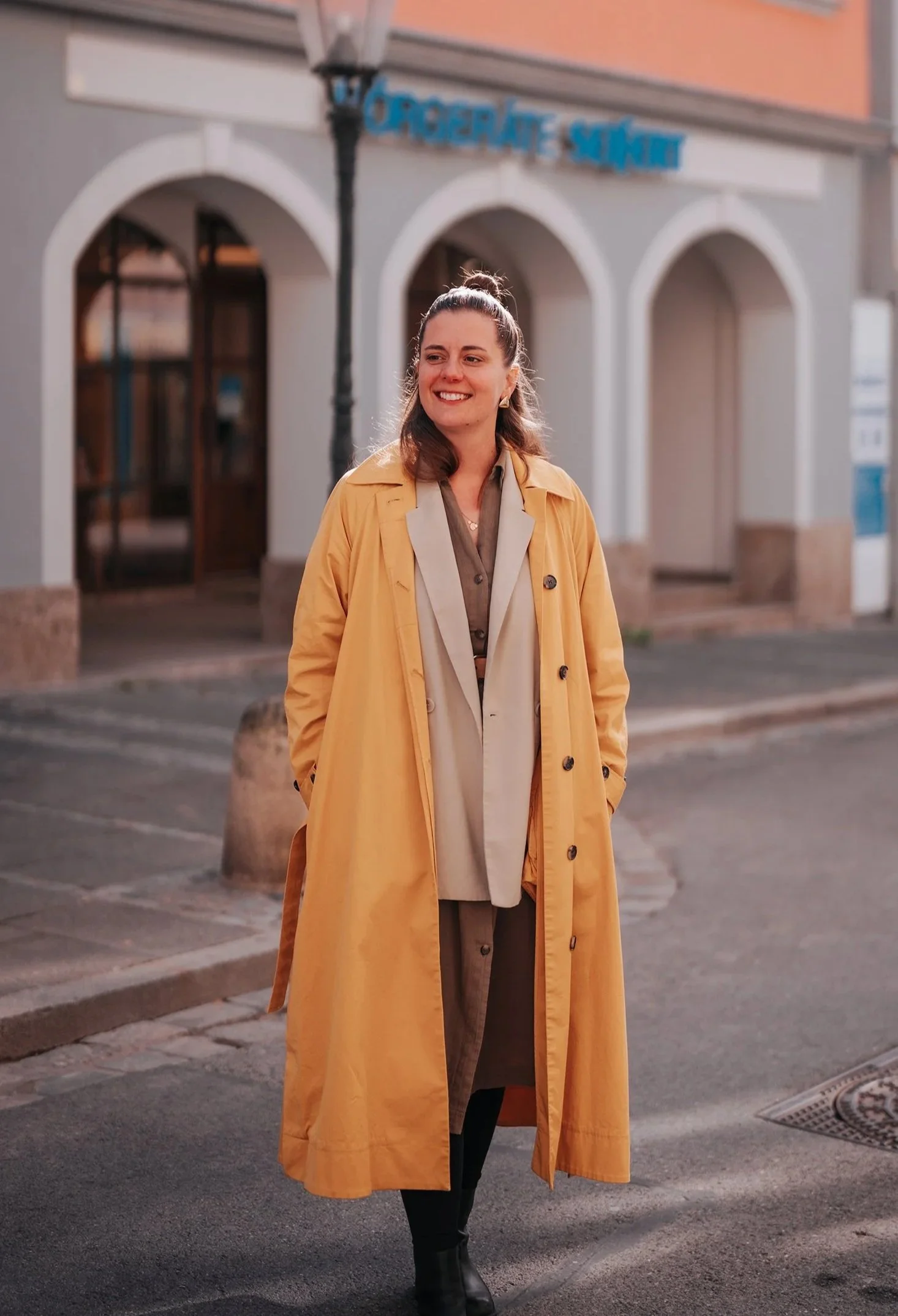 Eine lachende Frau in einem gelben Mantel, der auf einer Straße steht, im Hintergrund ein Gebäude mit einem Schild, das 'Grocery Markt' sagt.