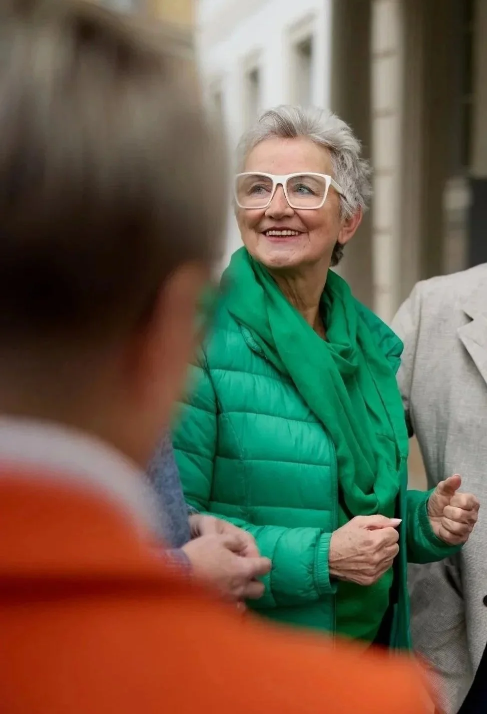 Eine ältere Frau mit kurzen grauen Haaren, weißen Brillen, grünem Pufferjacke und grüner Halskette, die lachend mit anderen Personen im Gespräch ist.