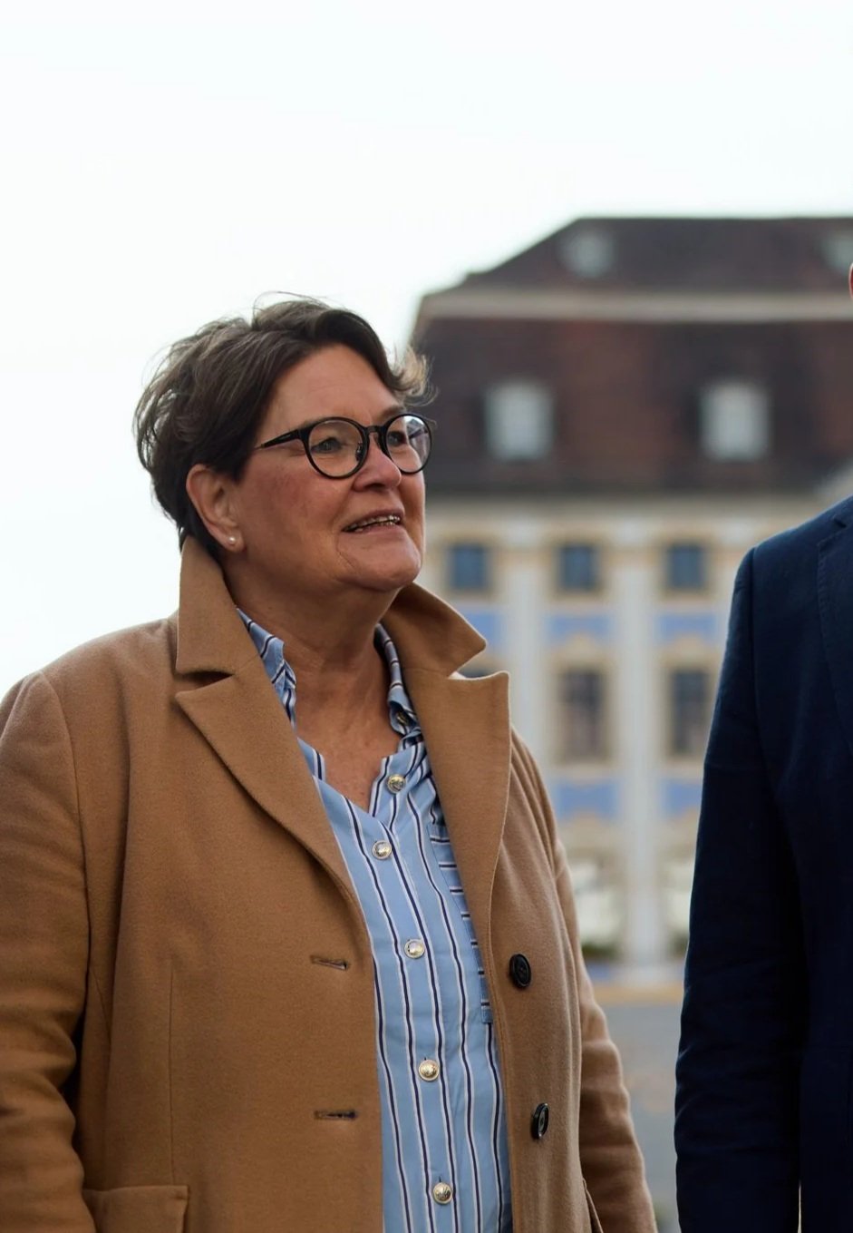 Eine ältere Frau mit kurzen braunen Haaren, Brille und einem beige Mantel, steht draußen vor einem Gebäude. Sie trägt eine blaue gestreifte Bluse.