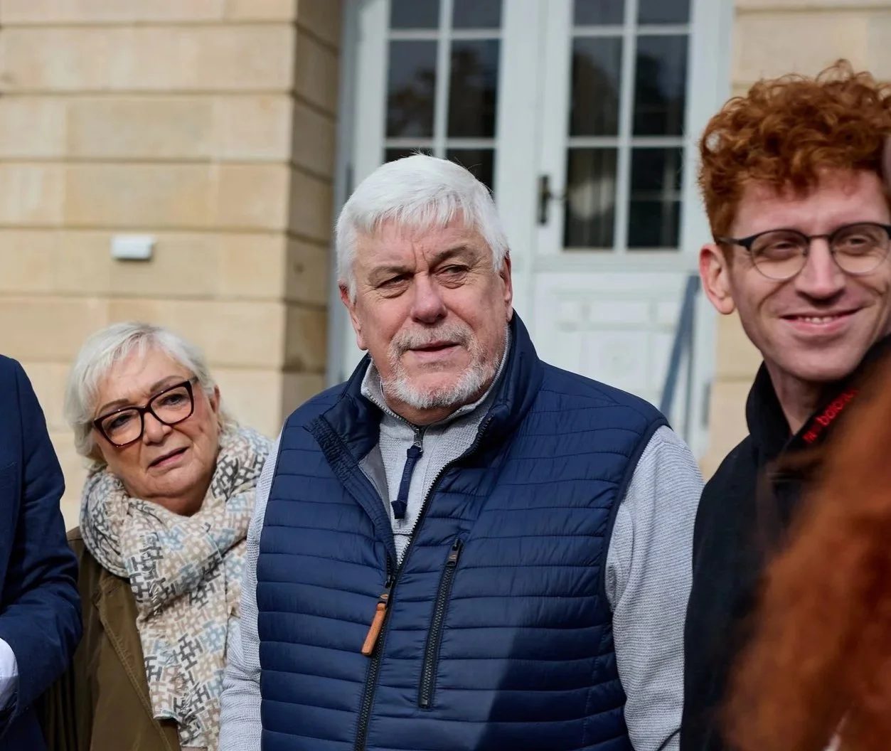Drei Menschen stehen vor einem Gebäude mit weißen Türen. Ein älterer Mann mit weißen Haaren und Bart trägt eine blaue wattierte Weste. Neben ihm steht eine ältere Frau mit kurz grauhaarigem Haar und Brille, die ein Halstuch trägt. Auf der rechten Sei