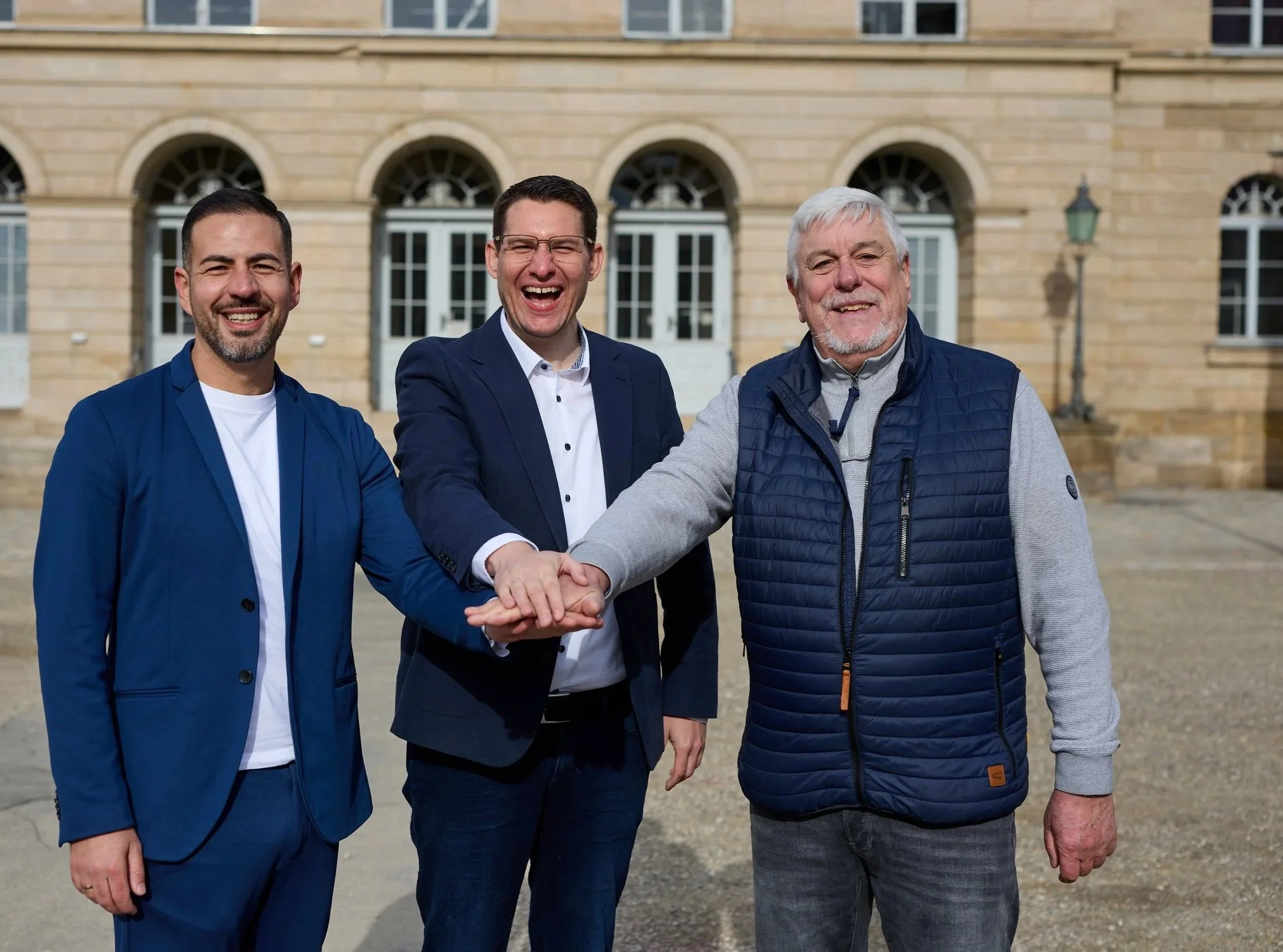 Drei Männer stehen vor einem historischen Gebäude, schütteln die Hände und lachen zusammen.