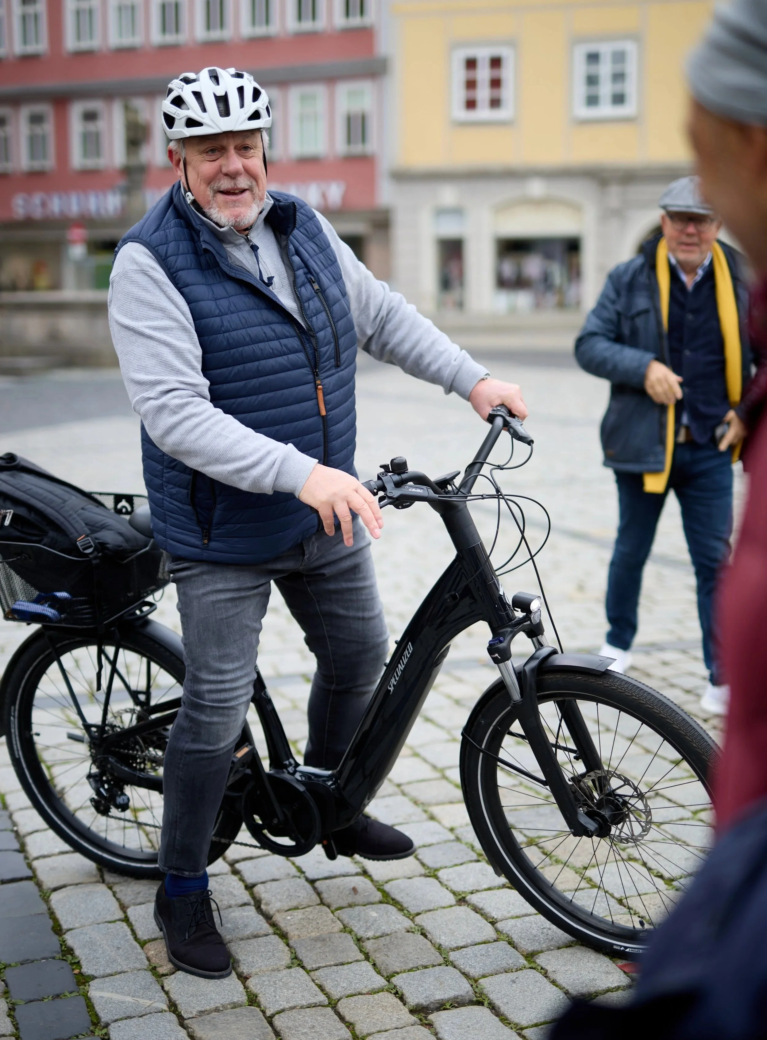 Ein älterer Mann steht auf einem Elektrofahrrad in einer Stadt, während andere Personen im Hintergrund stehen und sprechen.