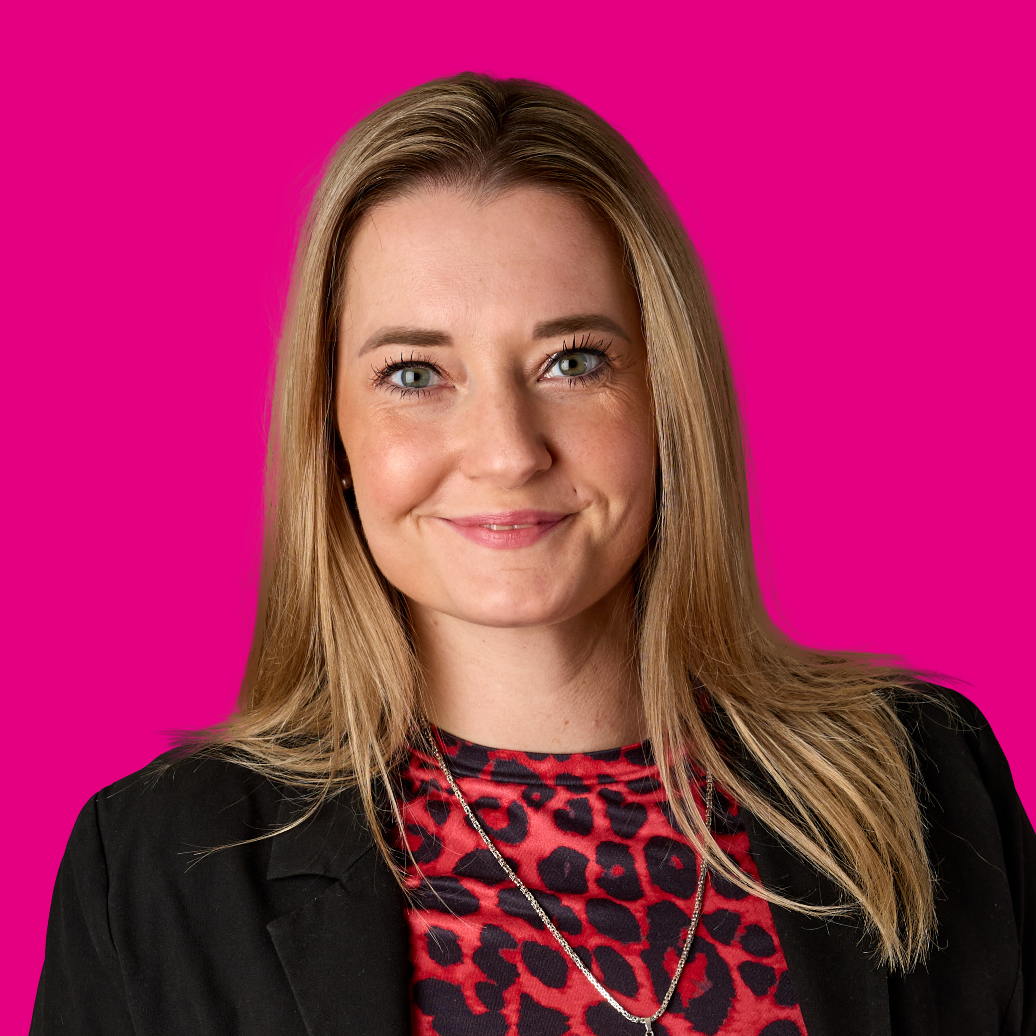 Porträt einer lächelnden Frau mit blonden Haaren gegen einen pinken Hintergrund, trägt ein rotes Top mit Schwarzfleckenmuster und eine schwarze Jacke.