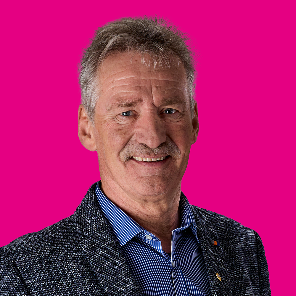 Ein älterer Mann mit grauem Haar und einem Schnurrbart, trägt eine blaue, gestreifte Hemd und eine dunkle Jacke, lächelt vor pinkem Hintergrund.