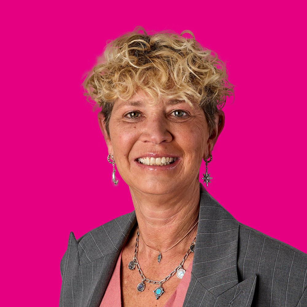 Eine lächelnde Frau mit kurzen, lockigen blonden Haaren trägt wellenförmige Ohrringe, mehrere Halsketten und ein graues Business-Outfit vor einem pinken Hintergrund.