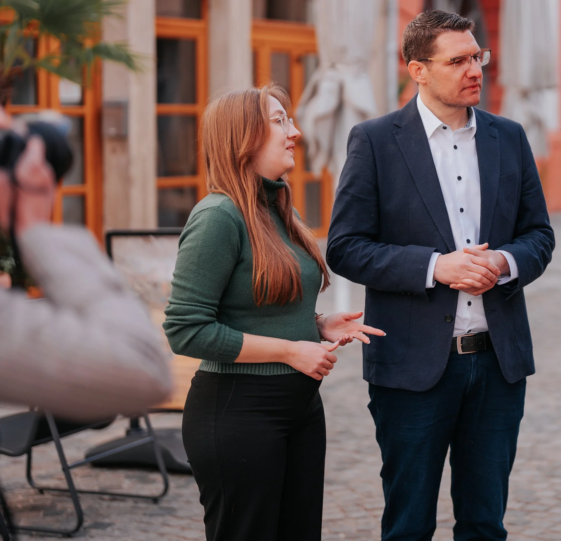 Zwei Menschen, eine junge Frau und ein Mann, sprechen in einem Außenbereich. Die Frau trägt eine grüne Pullover und schwarze Hosen, der Mann trägt ein dunkelblauer Anzug mit weißem Hemd. Im Hintergrund sind Fenster und Sträucher sichtbar.