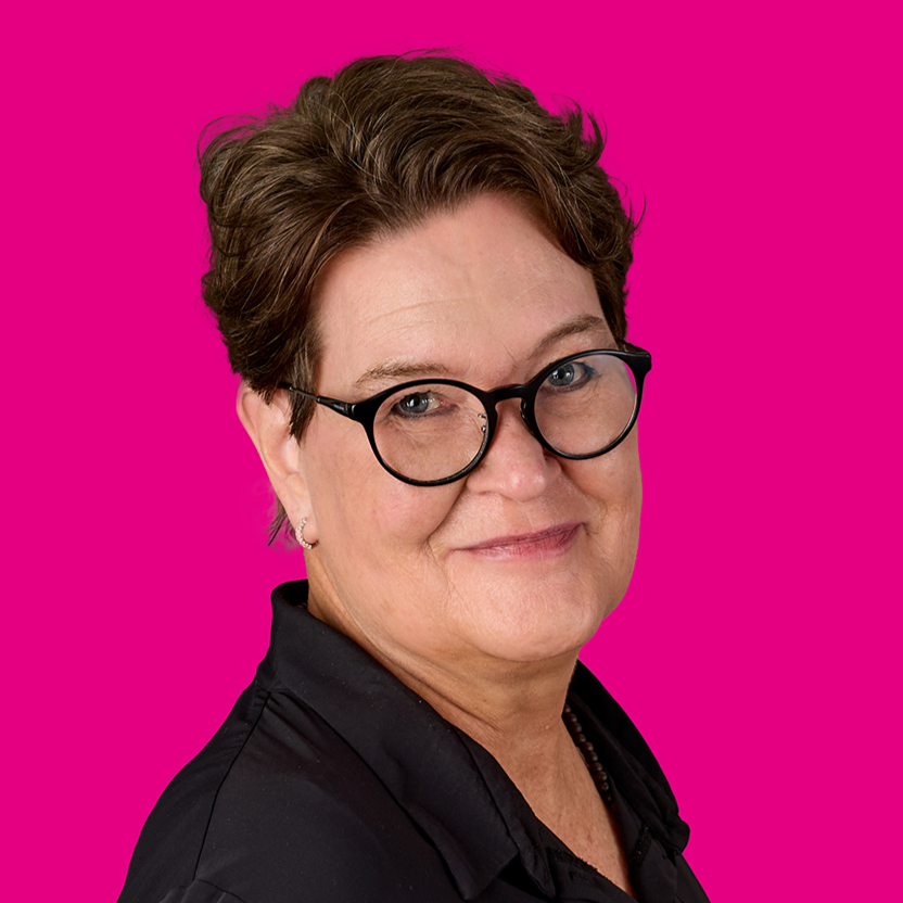 Porträt einer Frau mit Kurzhaarschnitt und Brille vor pinkem Hintergrund, lächelnd und in schwarzer Bluse.