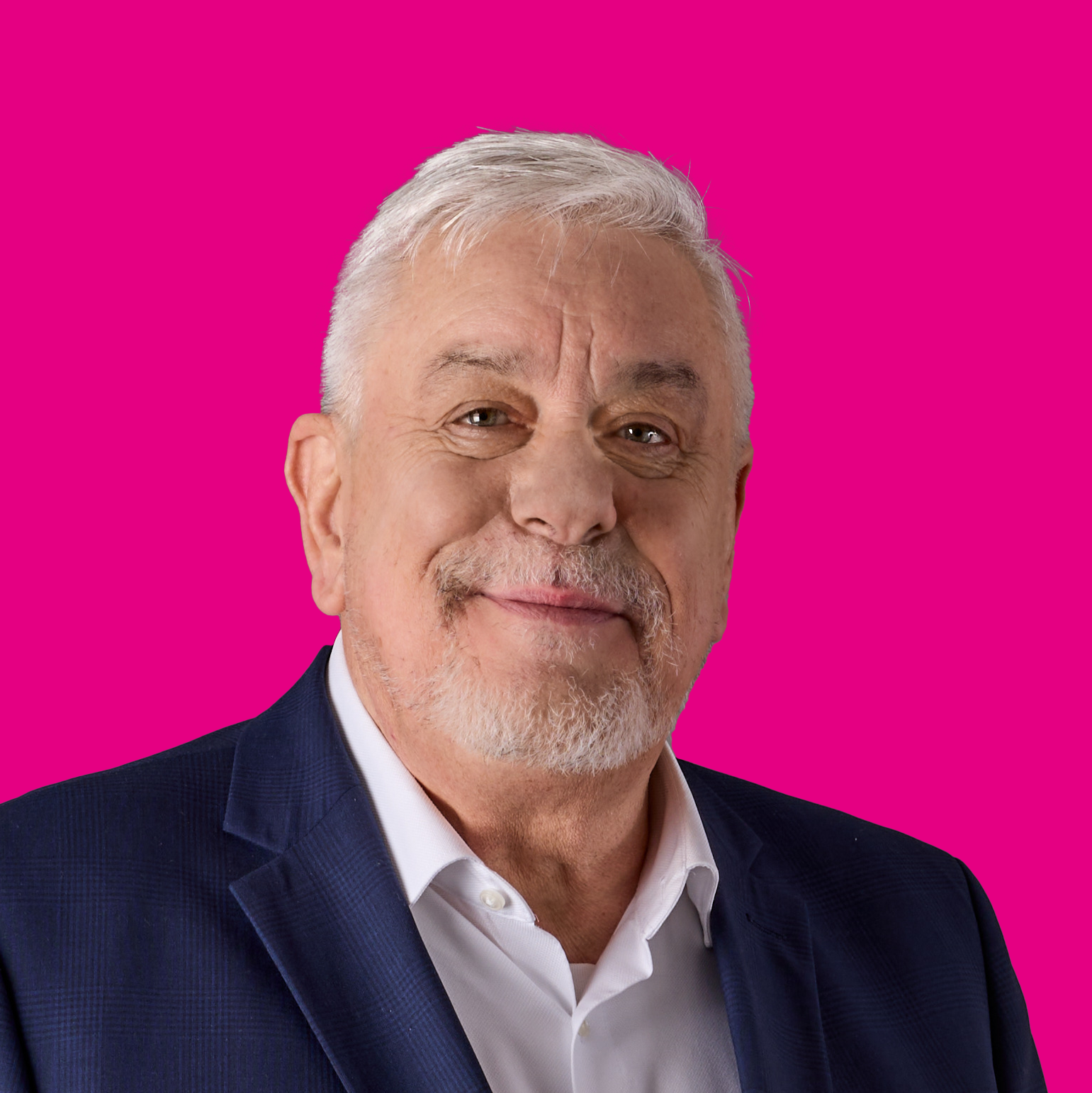 Nahaufnahme eines älteren Mannes mit weißem Haar, Bart, in dunklem Anzug und hellblauem Hemd vor pinkem Hintergrund.
