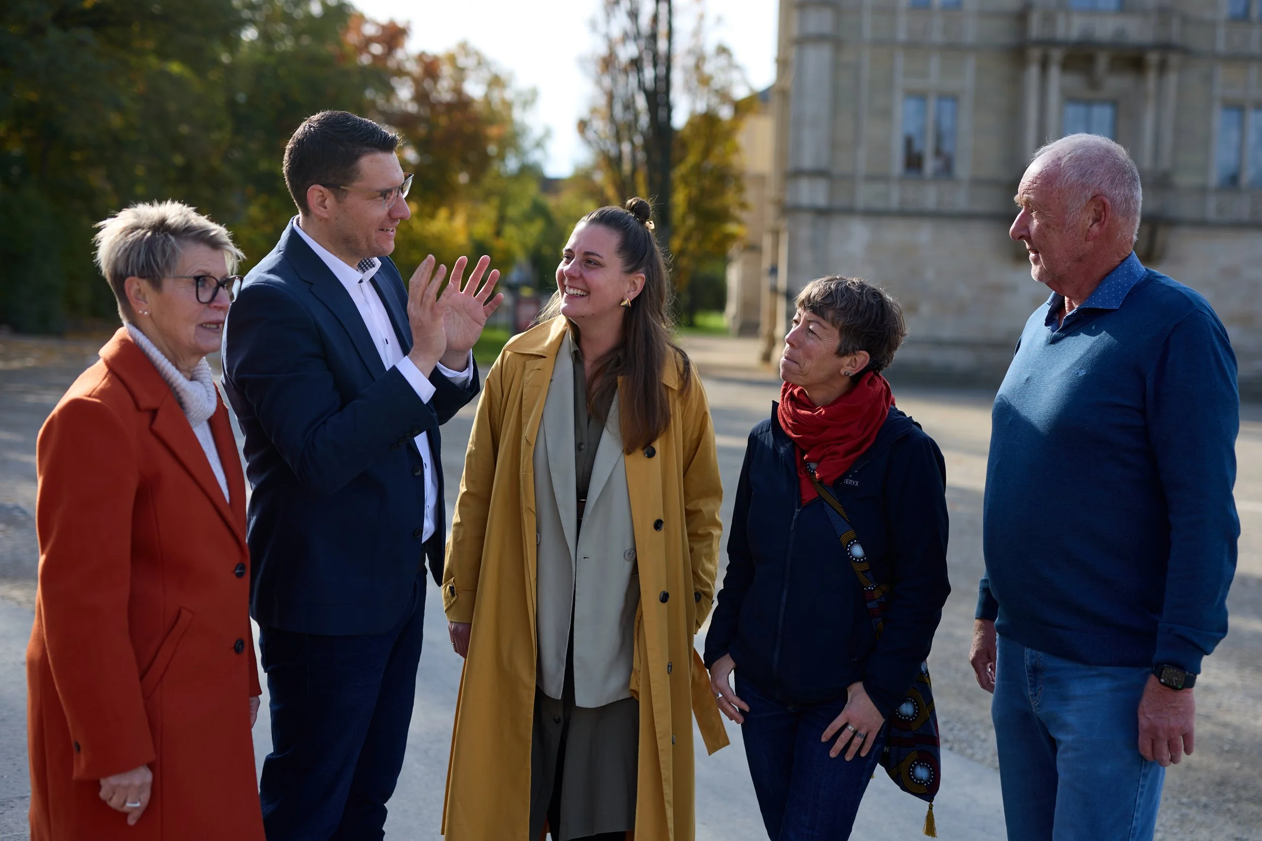 Fünf Menschen stehen im Gespräch vor einem historischen Gebäude in Herbstfarben. Die Stimmung ist freundlich und lebhaft.
