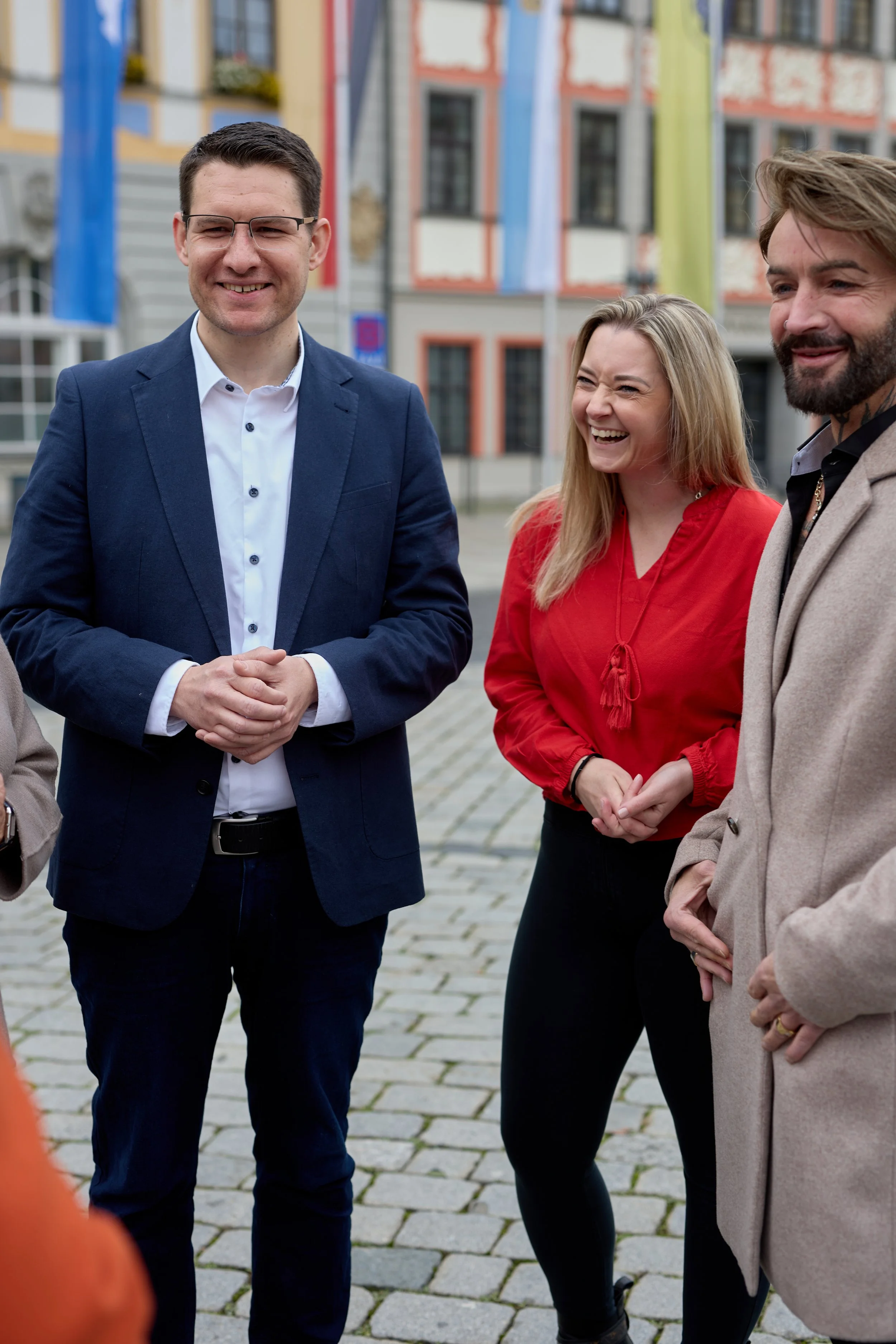 Vier Personen im Freien, die sich unterhalten und lachen, vor bunten Gebäuden und Flaggen.