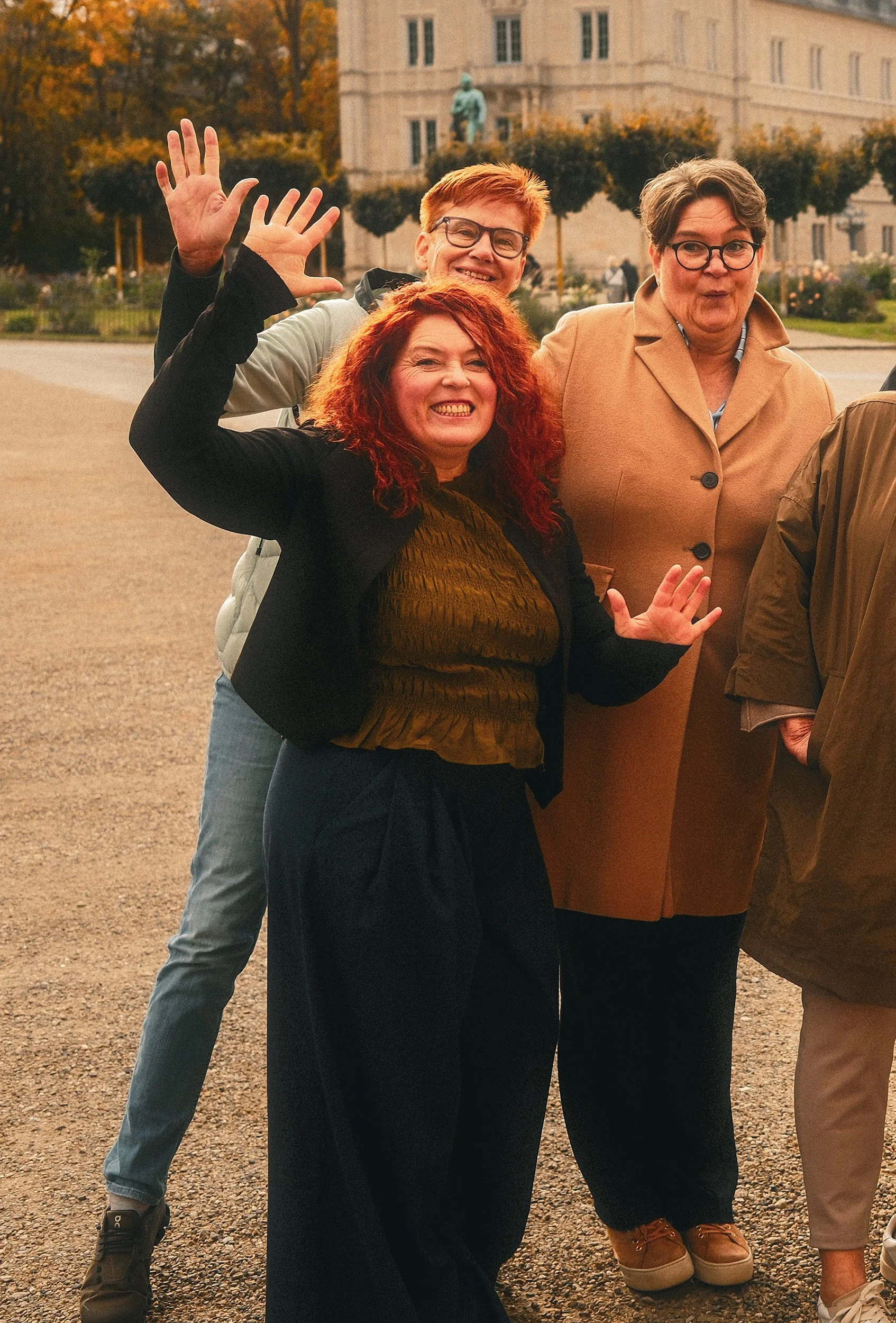 Drei lachende Frauen stehen im Freien, winken und posieren für die Kamera, im Hintergrund ein historisches Gebäude und Bäume mit Herbstlaub.