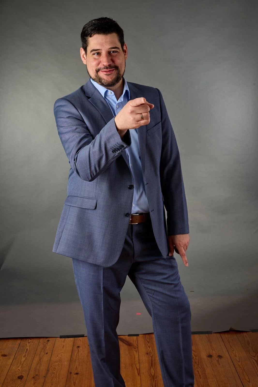 Ein Mann in einem dunklen Anzug, blauer Hemd, steht vor einem grauen Hintergrund, zeigt mit Finger auf die Kamera und lächelt.